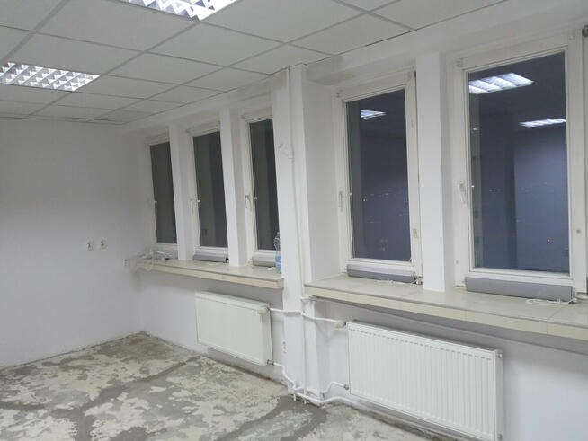 Pusty pokój biurowy przed remontem z białymi ścianami, grzejnikami i kilkoma oknami z widokiem na nocne miasto. Podłoga betonowa z widocznymi śladami po poprzednich pracach.