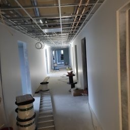 Korytarz w trakcie malowania na biało, widoczne rusztowania sufitowe i materiały budowlane na podłodze.