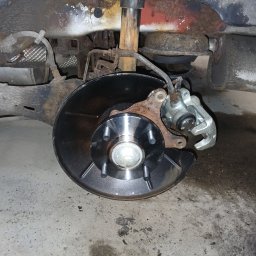 Car Serwis Sławno - Nowa tarcza hamulcowa z zaciskiem i elementami zawieszenia w samochodzie, widoczne ślady korozji na podwoziu.