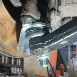 Wymiana sworznia wahacza w samochodzie w Sławnie, widoczny nowy element z niebieskim smarem, tarcza hamulcowa z widoczną korozją i skrzynka narzędziowa w tle.