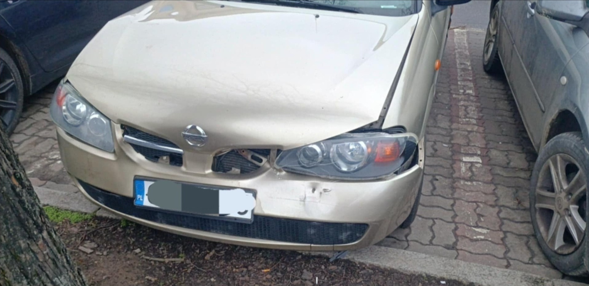 Złoty samochód Nissan Almera z uszkodzonym przodem, wgniecione błotniki i maska, pęknięty reflektor, widoczny fragment chłodnicy, zaparkowany na chodniku