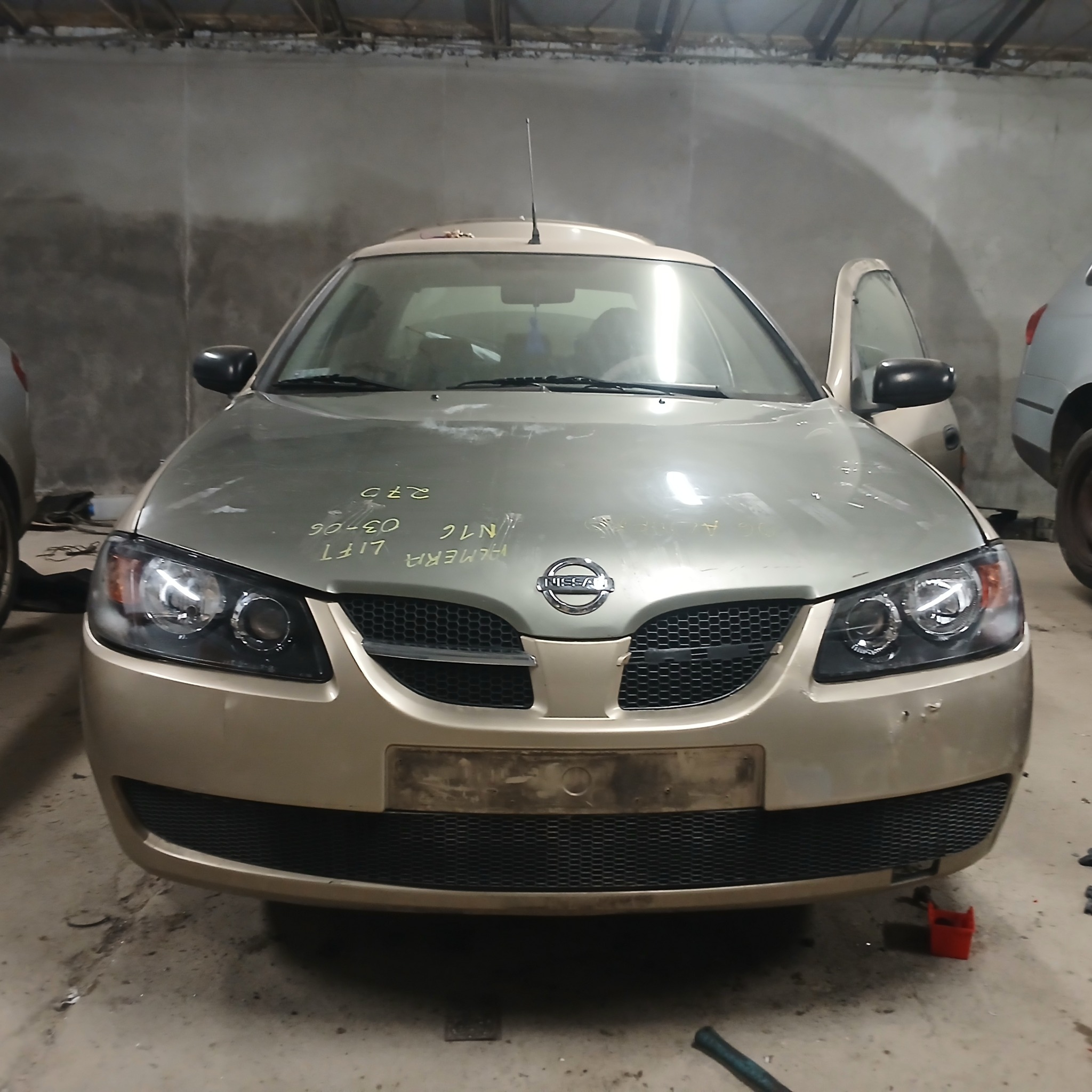 Przód samochodu Nissan Almera w warsztacie, z widocznymi notatkami na masce i otwartymi drzwiami pasażera.