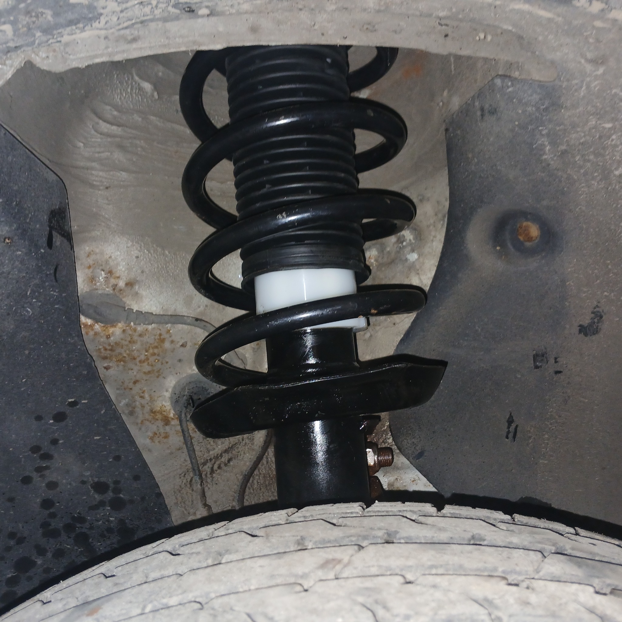 Czarny amortyzator sprężynowy z białym elementem mocującym, widoczny od dołu, z oponą w tle, w brudnym nadkolu z rdzawymi nalotami.