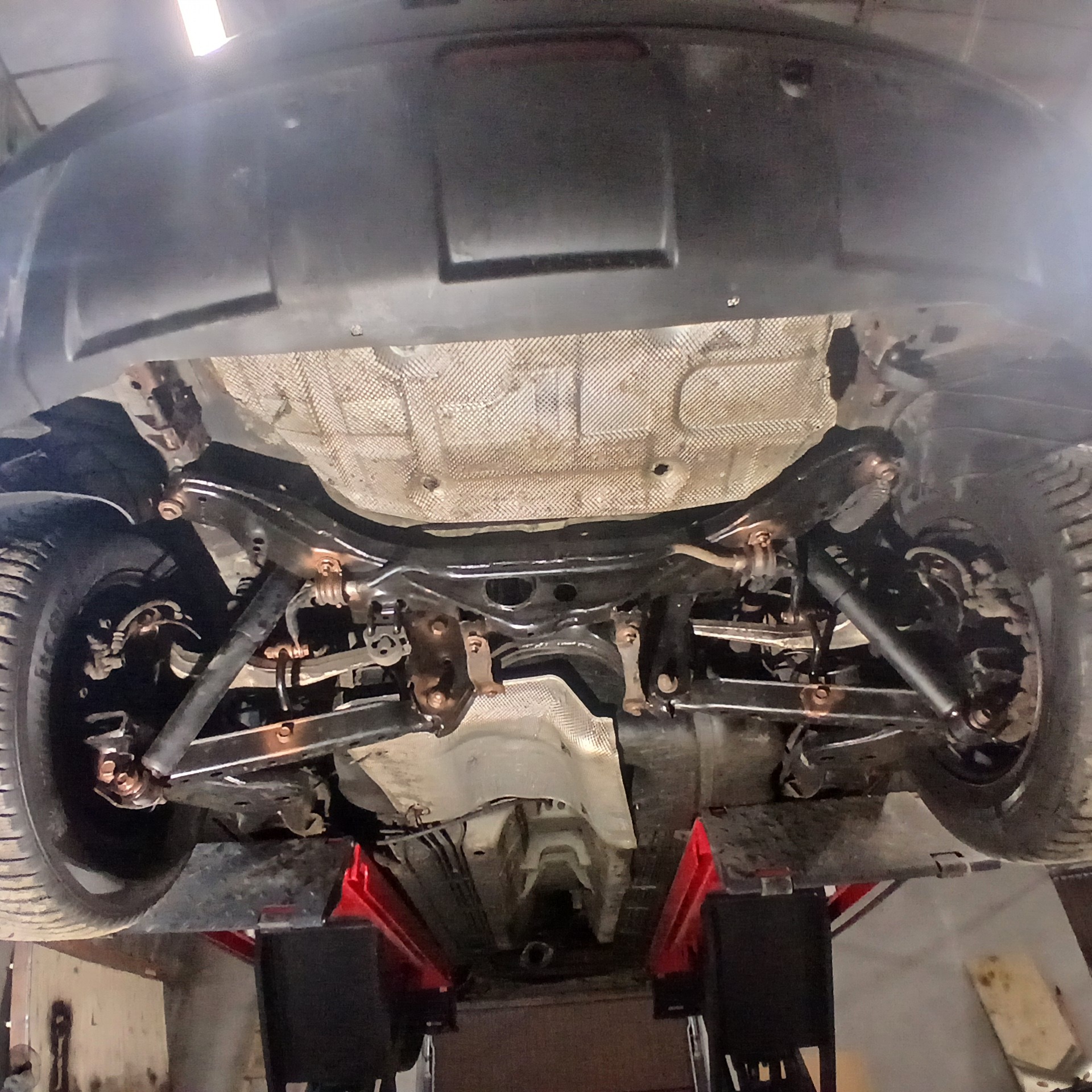 Widok podwozia samochodu na podnośniku warsztatowym, z widocznymi elementami zawieszenia, układu wydechowego oraz osłoną termiczną.