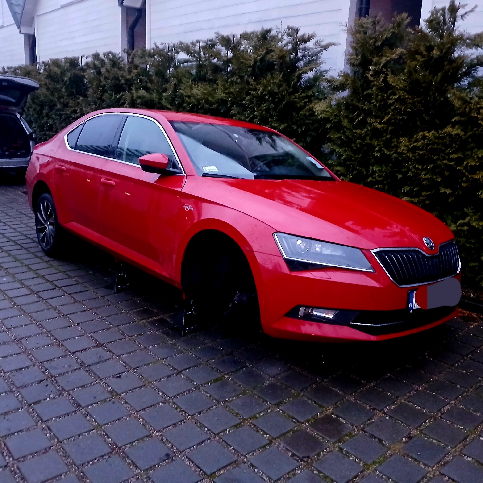 Czerwona Skoda Superb na podnośnikach, bez przedniego koła, na mokrej kostce brukowej, z bagażnikiem otwartym w tle, z widocznym fragmentem tablicy rejestracyjnej.