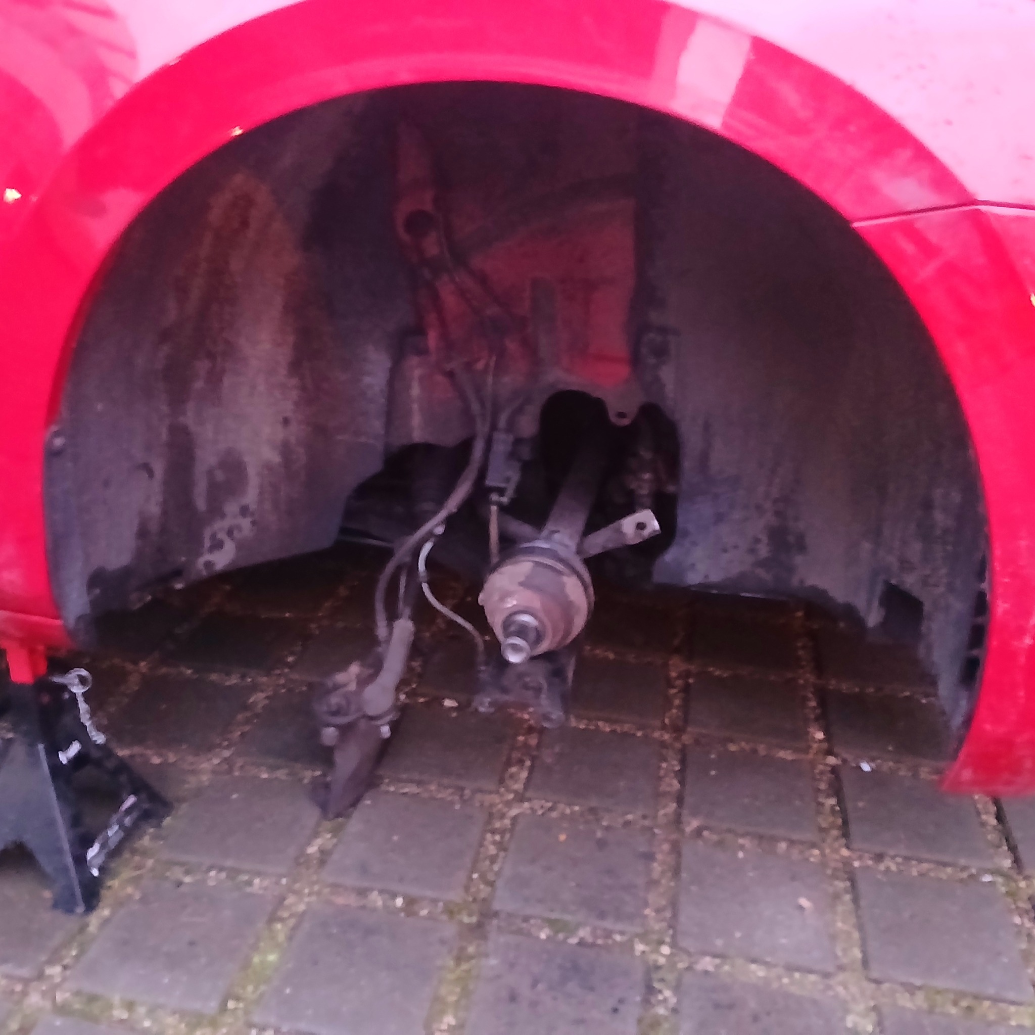 Czerwony samochód na podnośniku z widocznym, zdemontowanym zawieszeniem i tarczą hamulcową, na brukowanym podłożu.