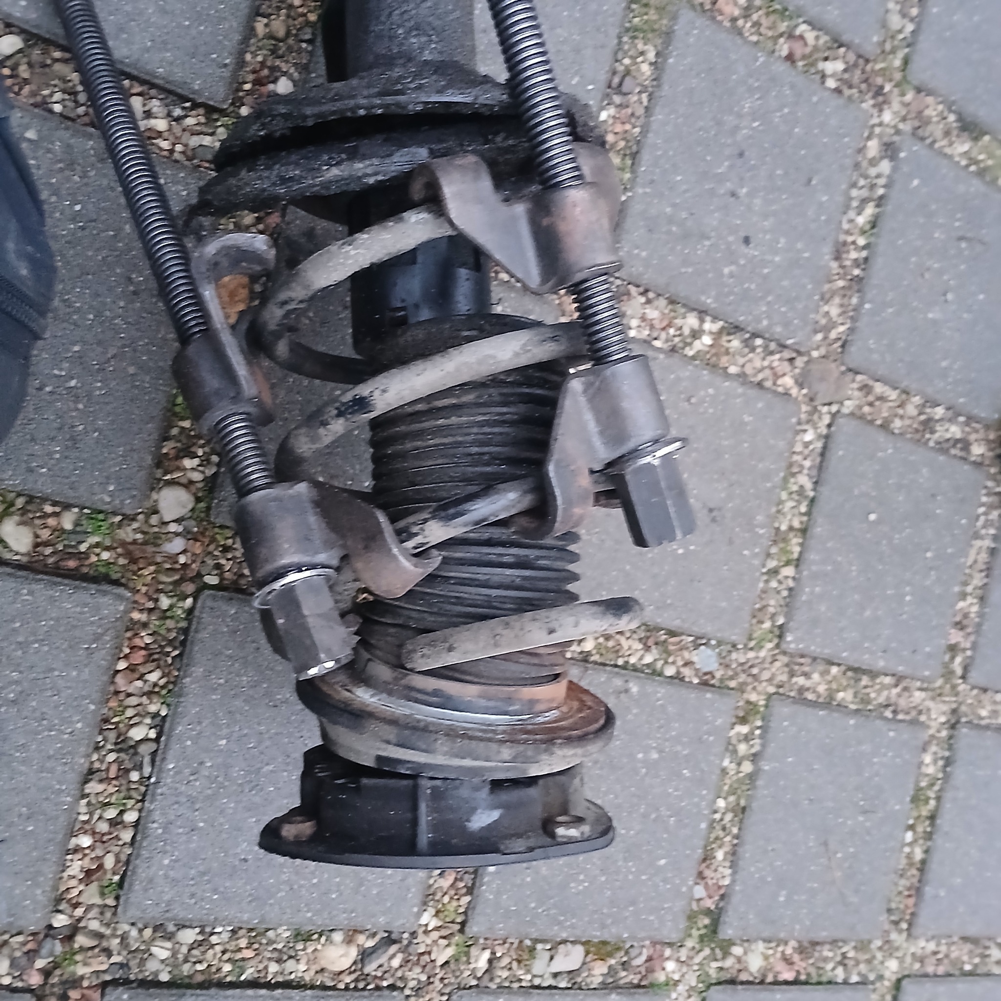 Sprężyna zawieszenia samochodowego ściśnięta specjalnymi ściskami, widoczna na tle betonowej kostki brukowej.