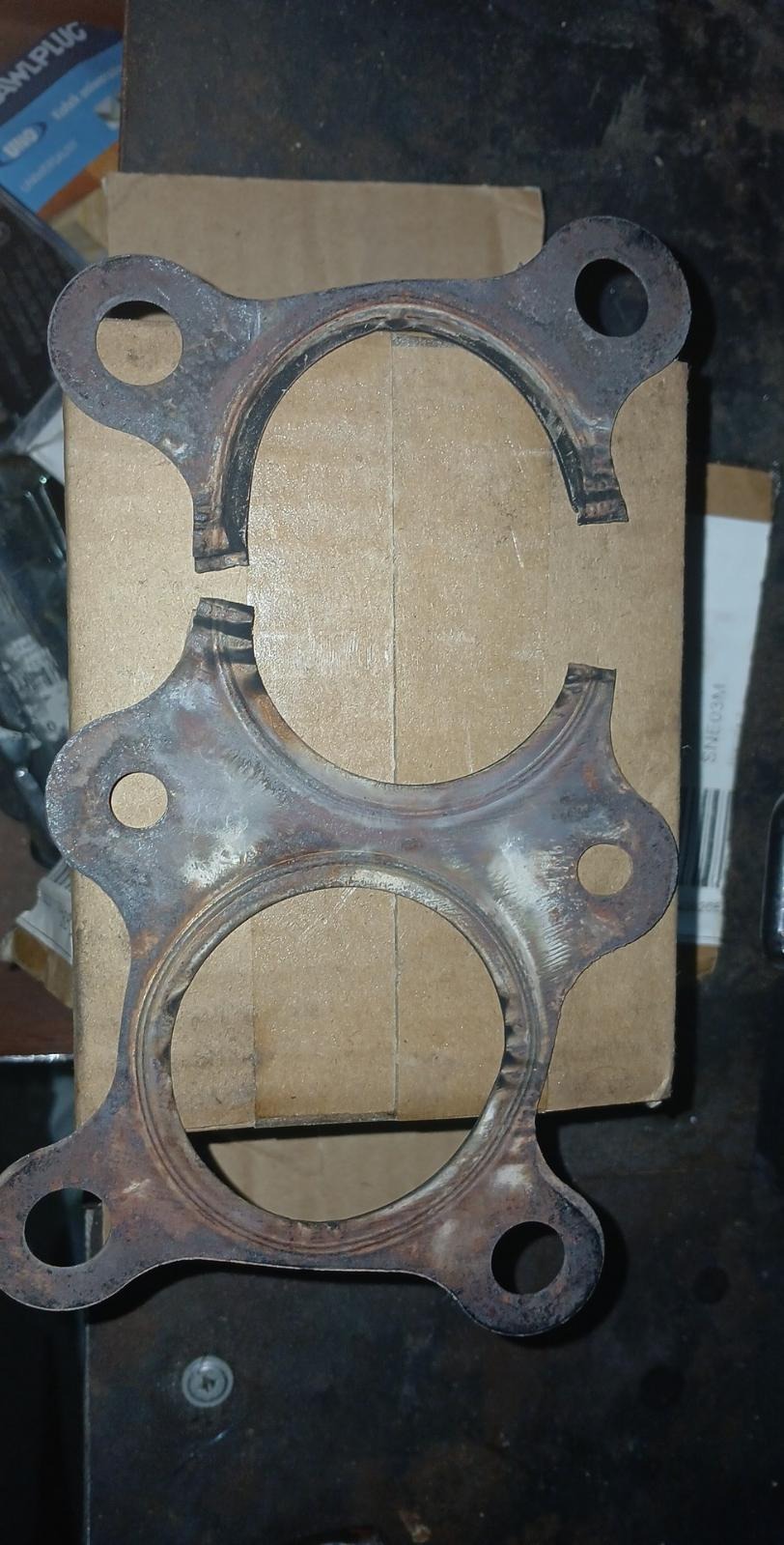 Zużyta, skorodowana uszczelka wydechu samochodowego, z widocznym pęknięciem, leżąca na kartonowym pudełku.