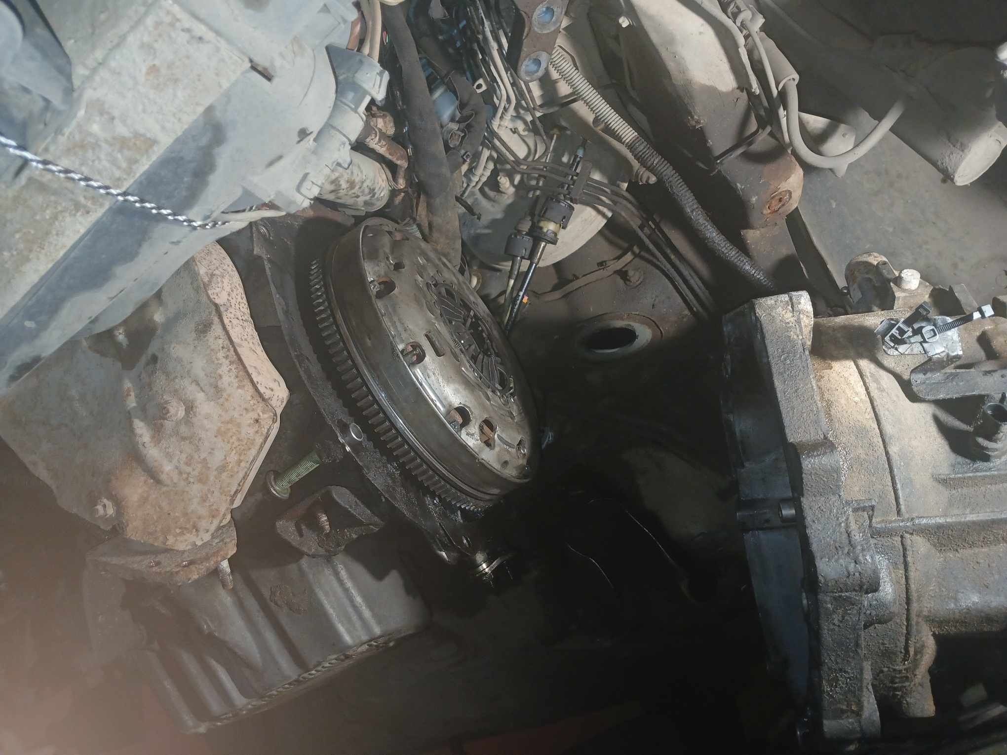 Widok od dołu na rozmontowany silnik samochodowy z widocznym sprzęgłem, skrzynią biegów i przewodami paliwowymi.