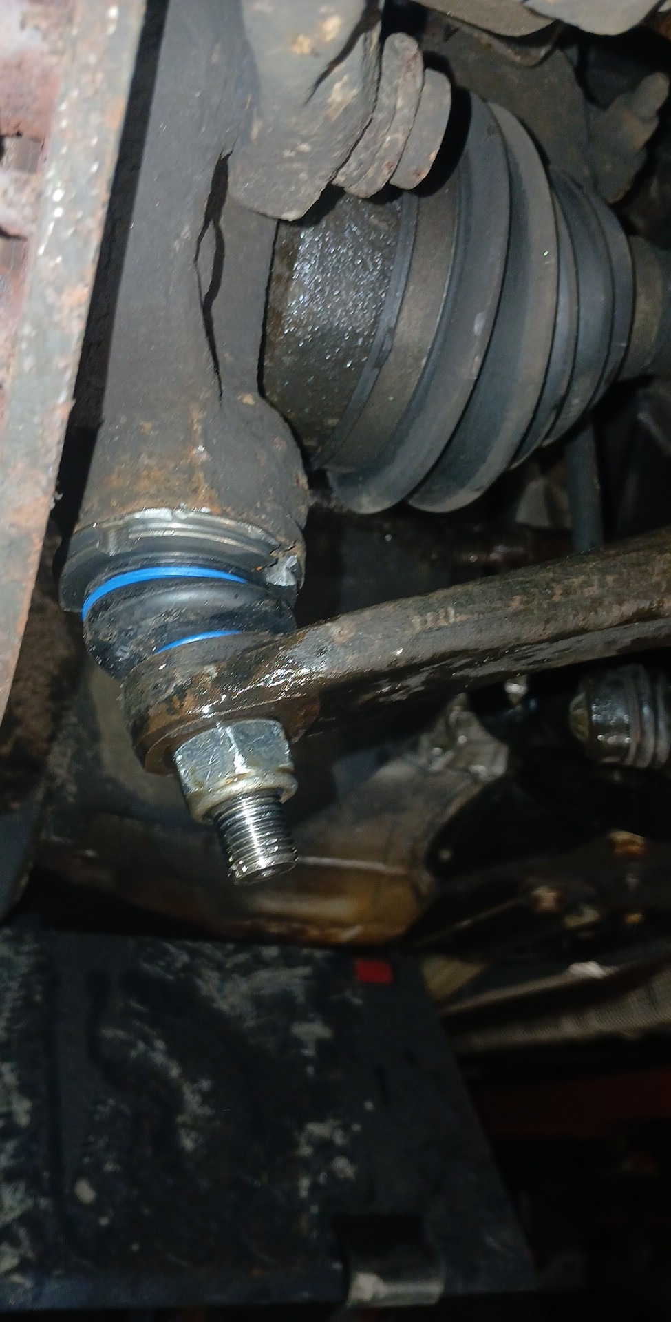 Element zawieszenia samochodu z widoczną tuleją z niebieskimi akcentami, nakrętką i przegubem, sfotografowany od spodu, z widocznymi śladami eksploatacji i zabrudzeniami.