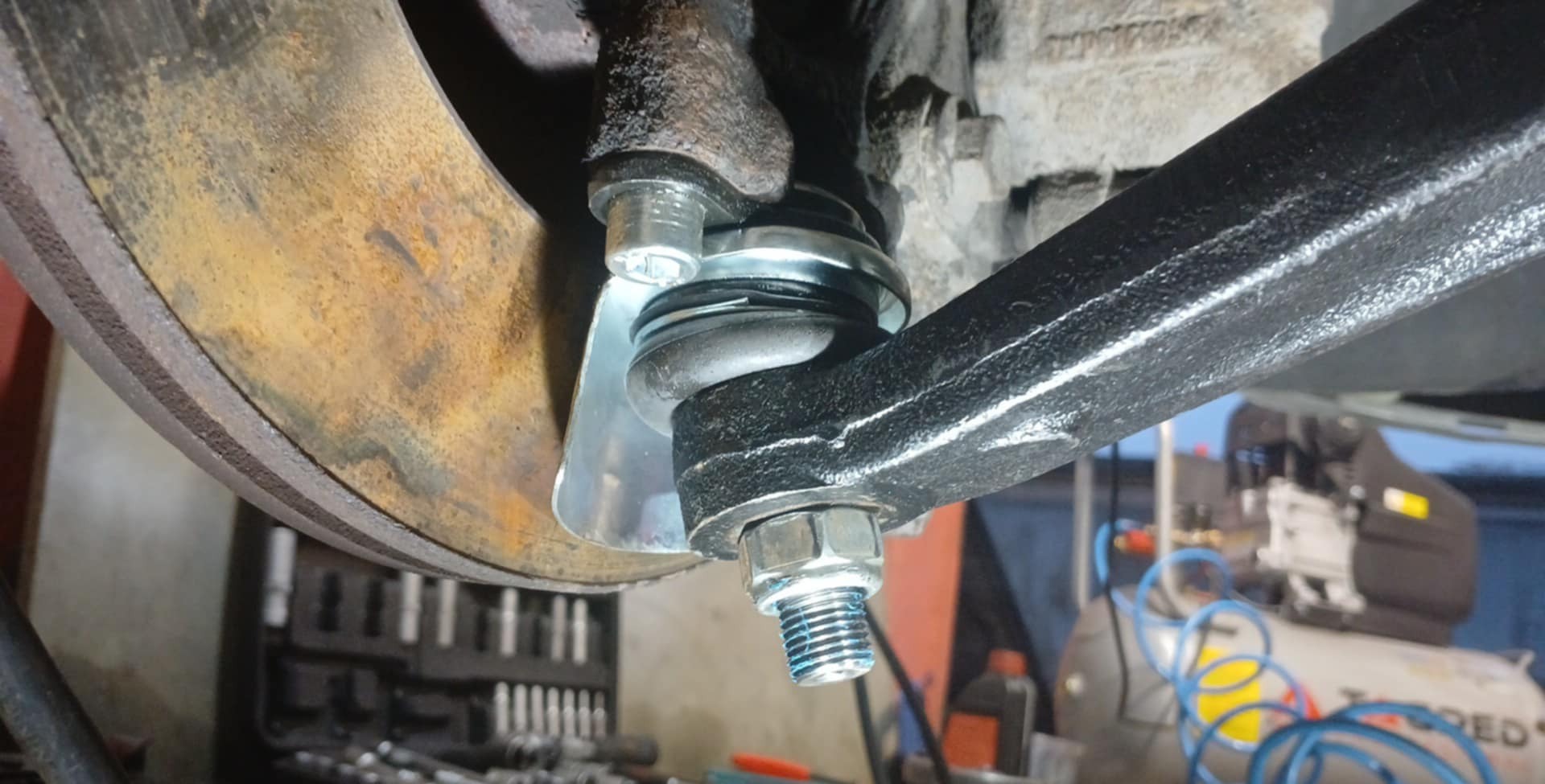 Wymiana sworznia wahacza w samochodzie w Sławnie, widoczny nowy element z niebieskim smarem, tarcza hamulcowa z widoczną korozją i skrzynka narzędziowa w tle.