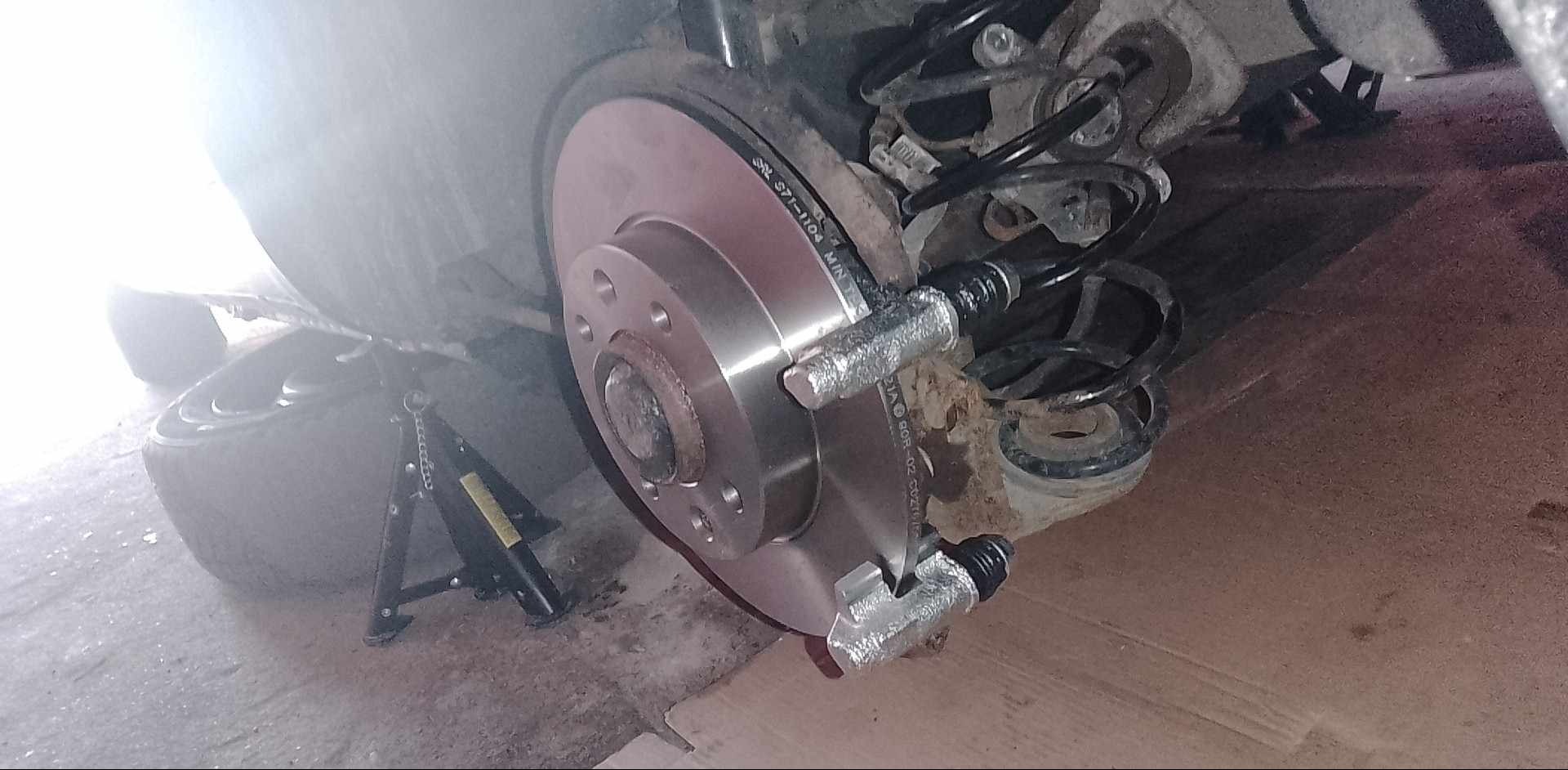 Nowa tarcza hamulcowa i zacisk zamontowane na samochodzie, widoczny podnośnik podtrzymujący pojazd, sprężyna zawieszenia w tle.