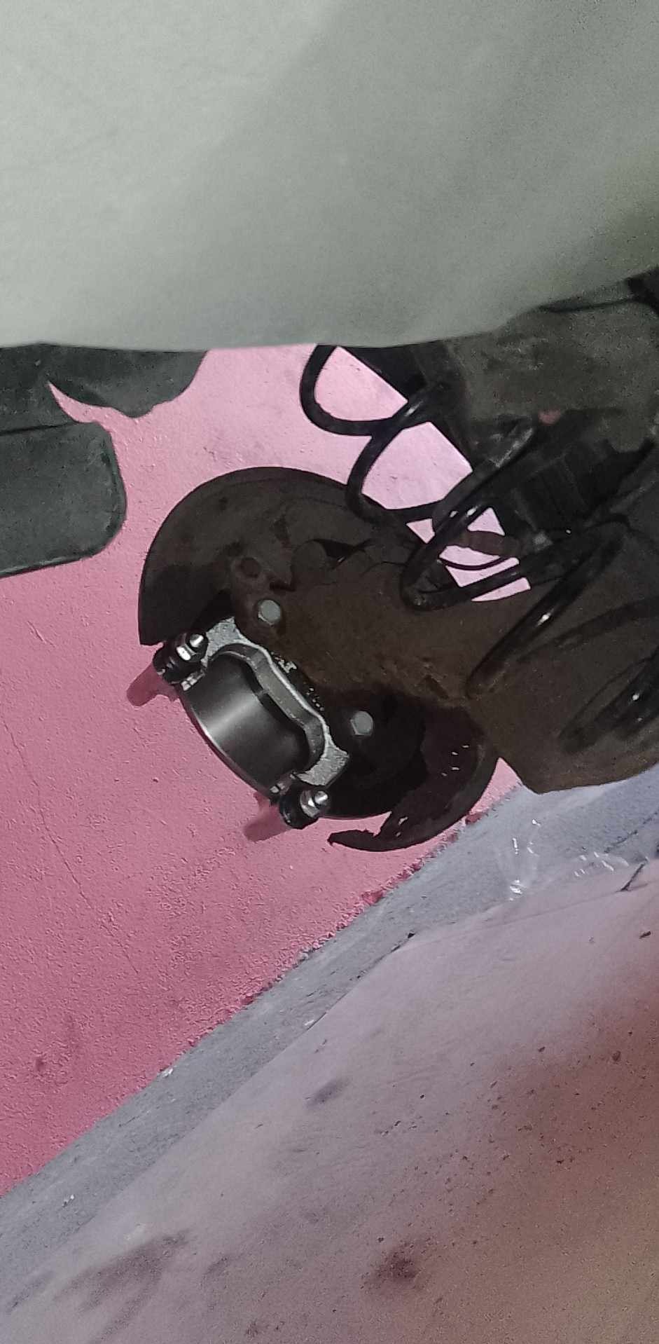 Fragment zawieszenia samochodowego z nowym elementem tulei, widoczne sprężyny i tarcza hamulcowa, tło różowe.