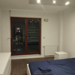 Mariusz Orczyk - Jasne wnętrze sypialni z parkietem, białymi ścianami, oknem balkonowym w brązowej ramie i widocznym odbiciem osoby. Minimalistyczny design, białe meble, niebieska pościel.