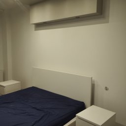 Mariusz Orczyk - Minimalistyczna sypialnia po malowaniu: białe ściany, łóżko z granatową pościelą, szafki nocne. Górne oświetlenie dodaje nowoczesnego charakteru.
