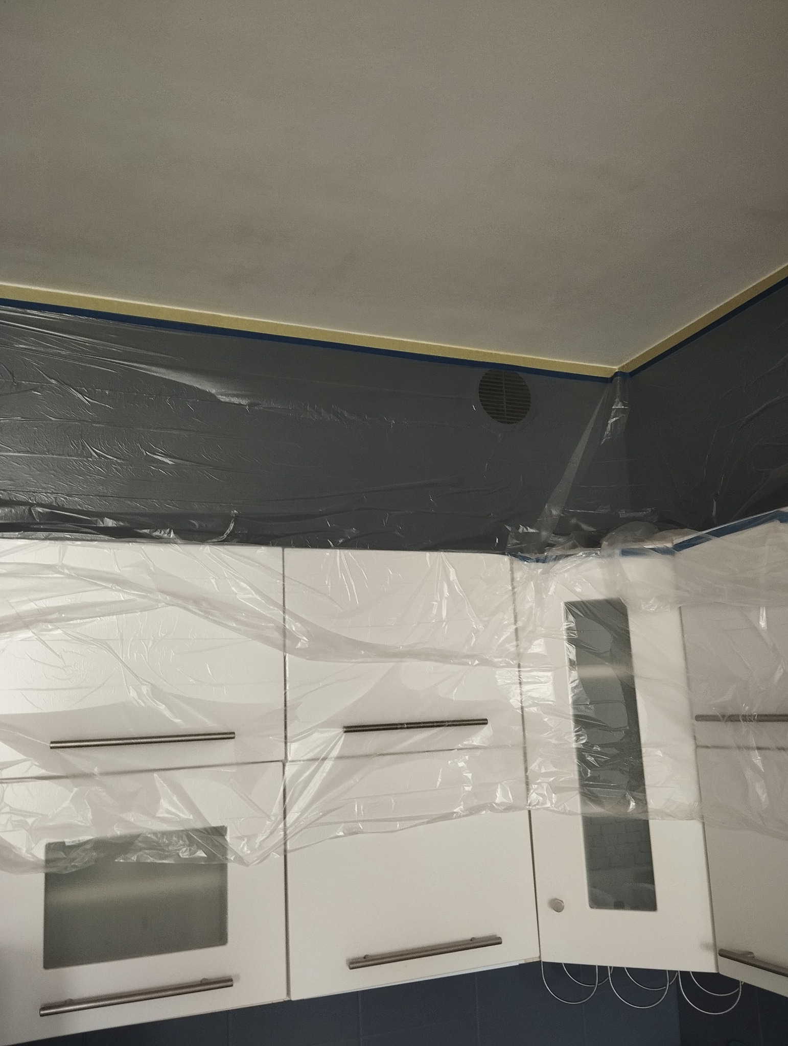 Przygotowanie do odświeżenia kuchni: Sufit i górne szafki zabezpieczone folią malarską, ściany częściowo pomalowane na szaro, taśma malarska oddziela sufit.