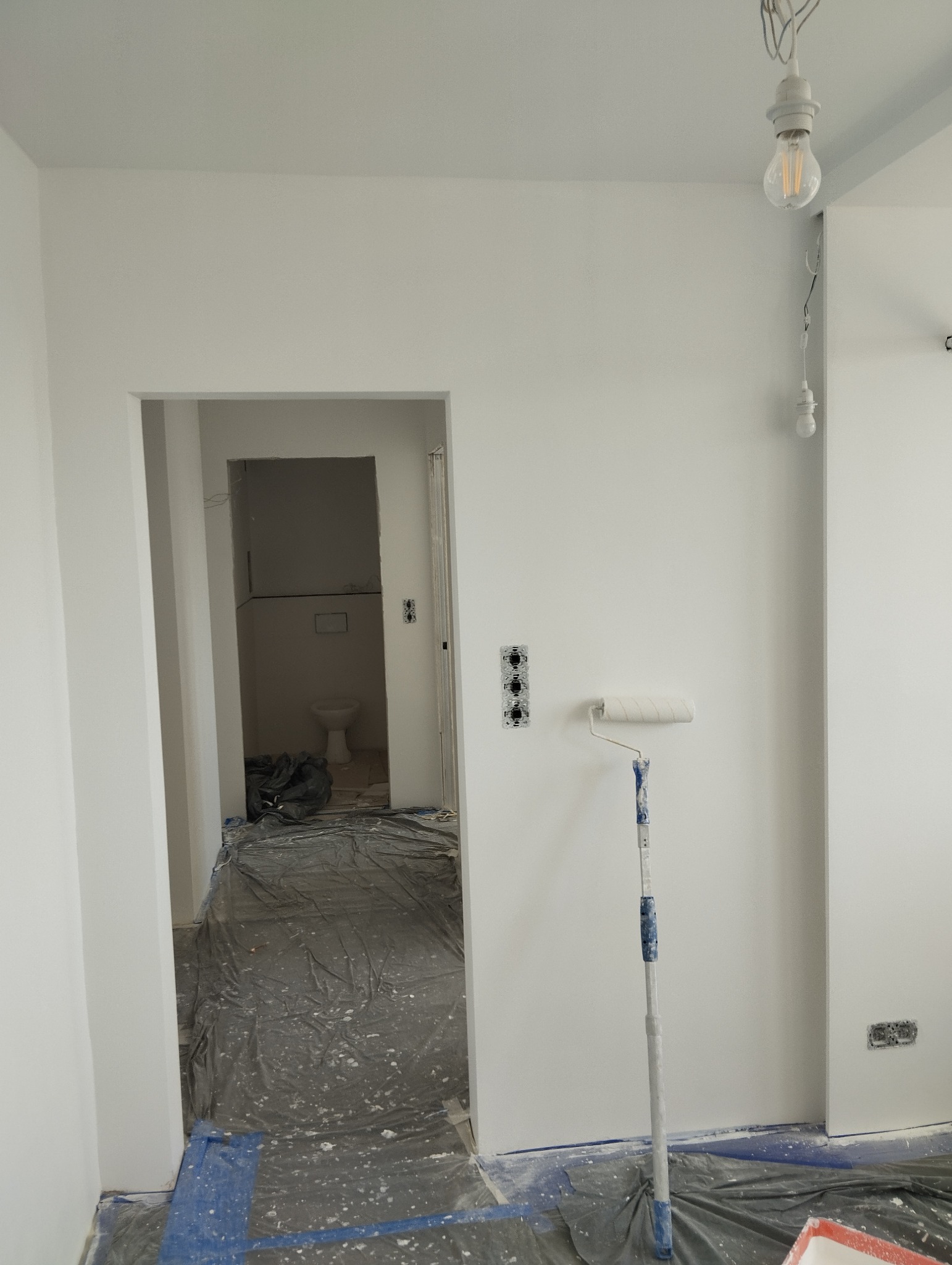 Białe ściany po malowaniu, widoczny wałek malarski, zabezpieczona folią podłoga i gniazdka elektryczne. W tle widoczne wejście do łazienki.