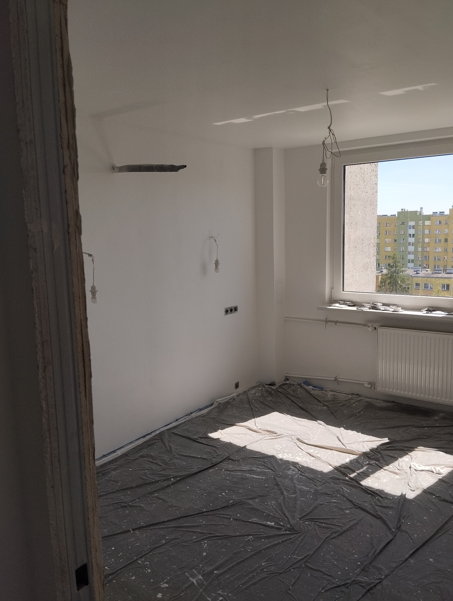 Białe ściany w pokoju po malowaniu, zabezpieczona podłoga folią malarską, widoczne gniazdka elektryczne i wiszące przewody oświetleniowe, okno z widokiem na budynki.