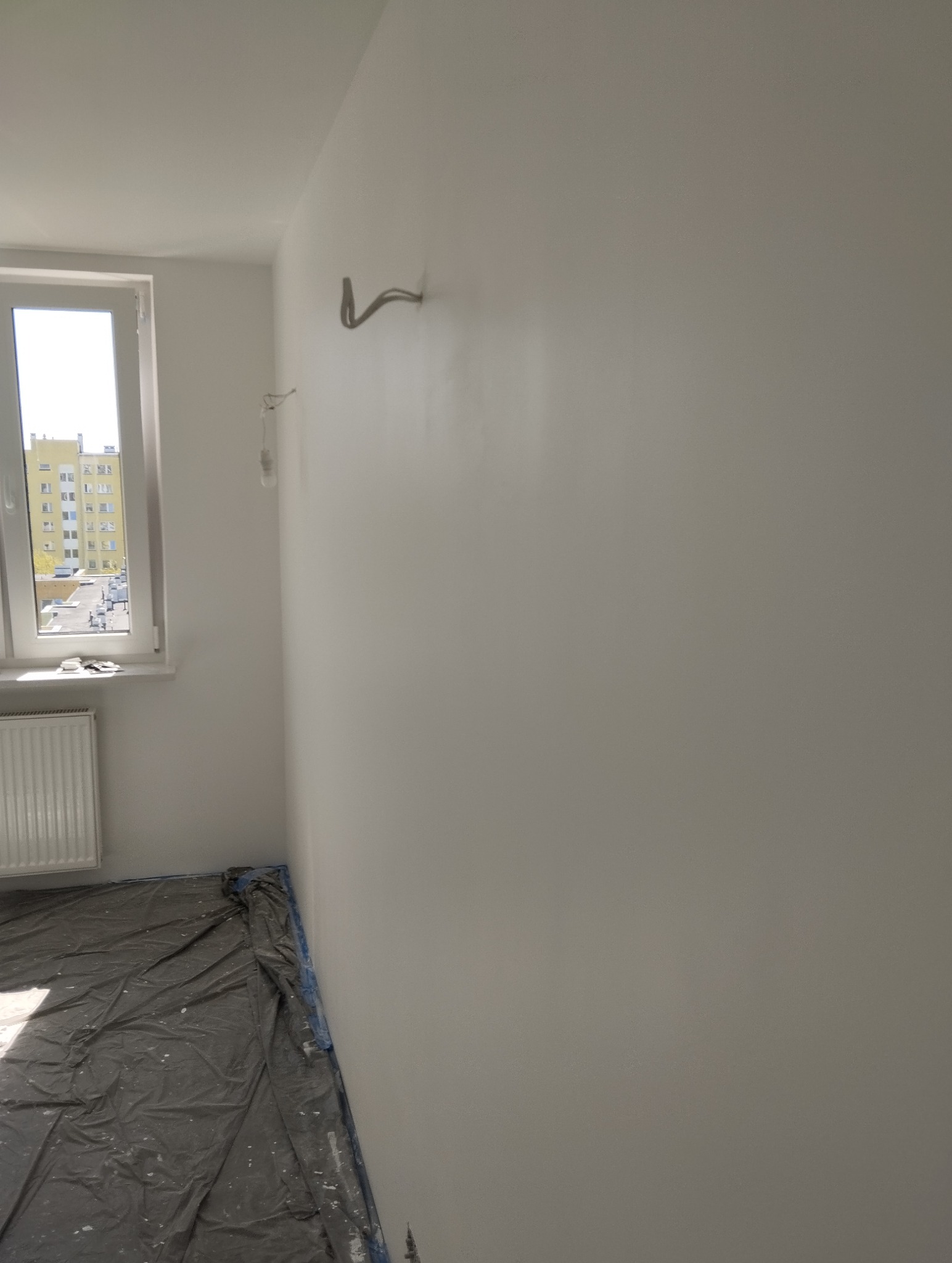 Świeżo pomalowane, jasne wnętrze z zakrytą podłogą. Widoczne wystające kable i rurki. Naturalne światło wpada przez okno.