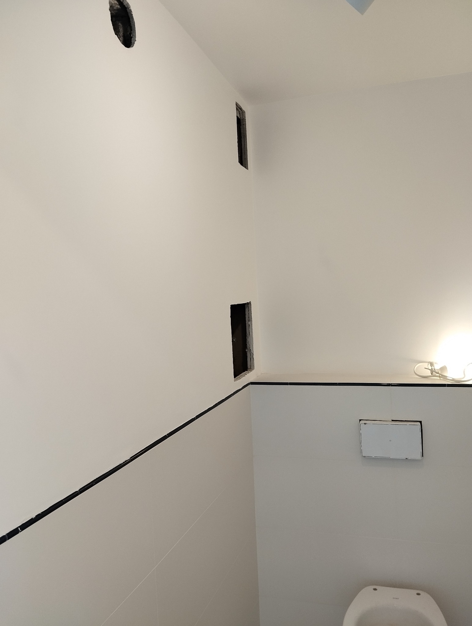 Biała ściana w łazience z otworem wentylacyjnym, dwoma otworami na instalacje i częściowo widoczną toaletą. Widoczne płytki z czarną listwą.