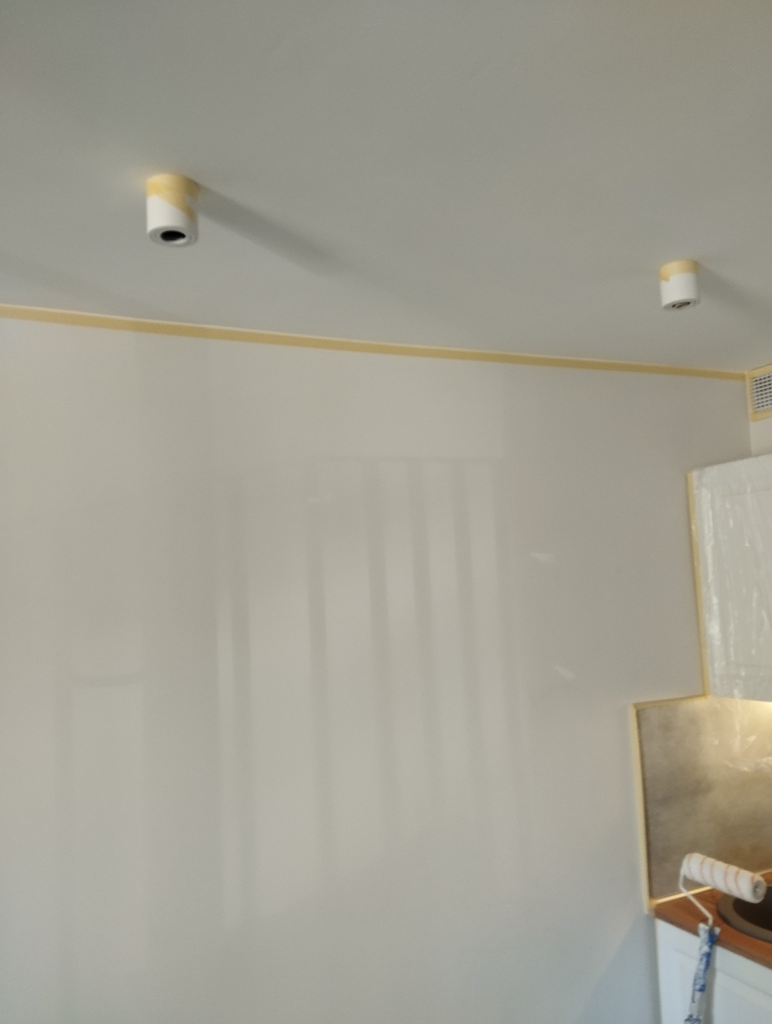 Świeżo pomalowana, jasna ściana z taśmą malarską przy suficie i rolką do malowania w dolnym rogu. Widoczne lampy sufitowe.