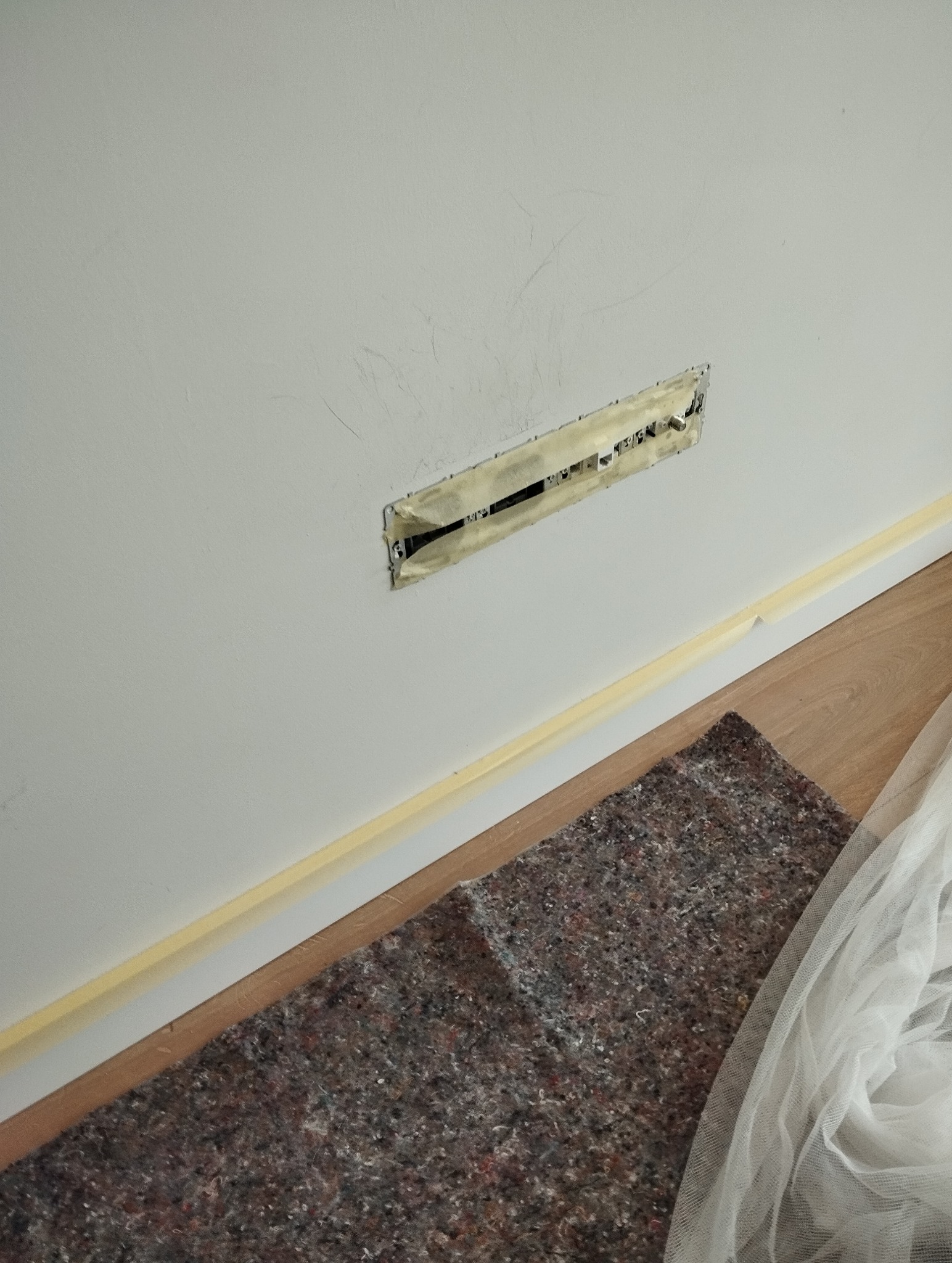 Ściana z otworem na instalację, zabezpieczonym taśmą malarską, przygotowana do dalszych prac wykończeniowych. Widoczna podłoga i materiał zabezpieczający.