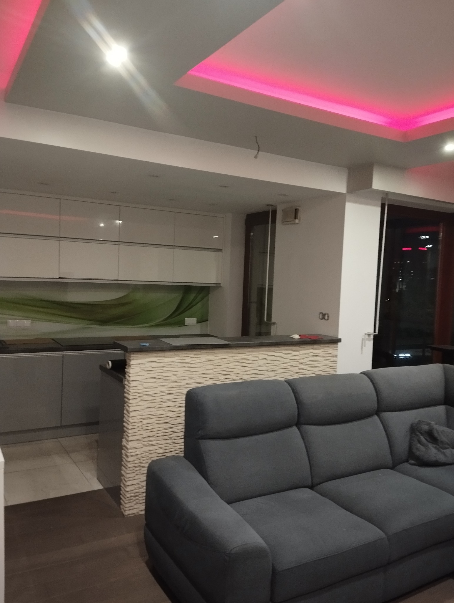 Nowoczesna kuchnia z białymi szafkami i zielonym panelem szklanym. Blat z mozaiką kamienną, oświetlenie LED sufitowe w kolorze różowym. Szary narożnik w salonie.