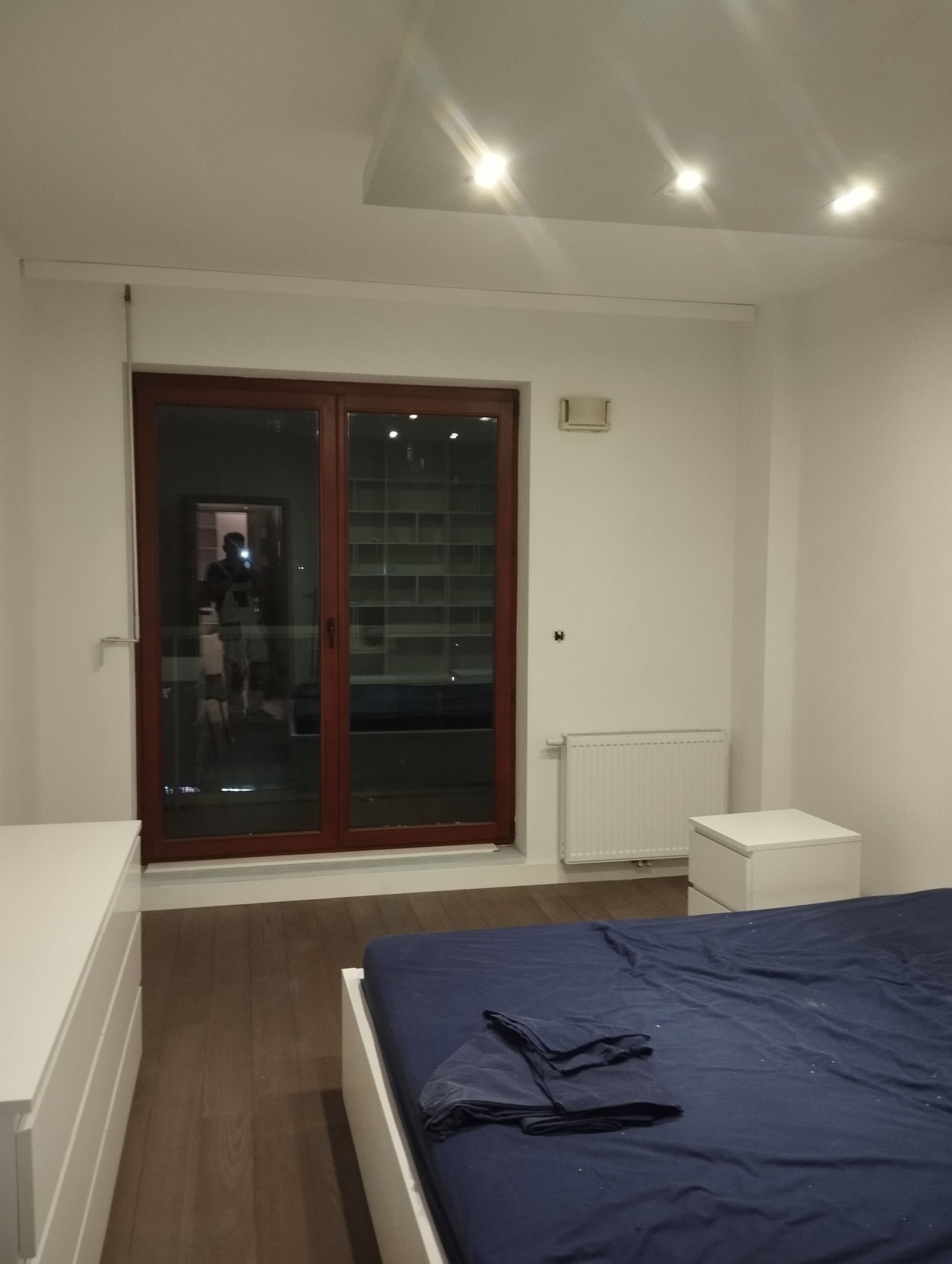 Jasne wnętrze sypialni z parkietem, białymi ścianami, oknem balkonowym w brązowej ramie i widocznym odbiciem osoby. Minimalistyczny design, białe meble, niebieska pościel.