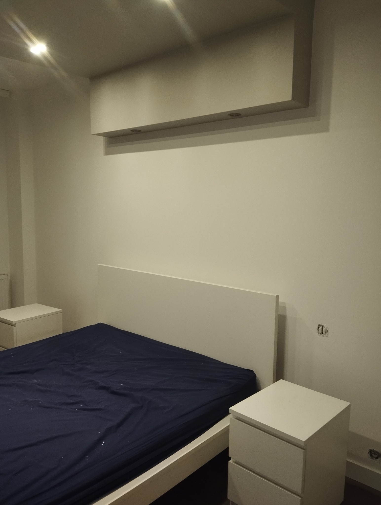 Minimalistyczna sypialnia po malowaniu: białe ściany, łóżko z granatową pościelą, szafki nocne. Górne oświetlenie dodaje nowoczesnego charakteru.