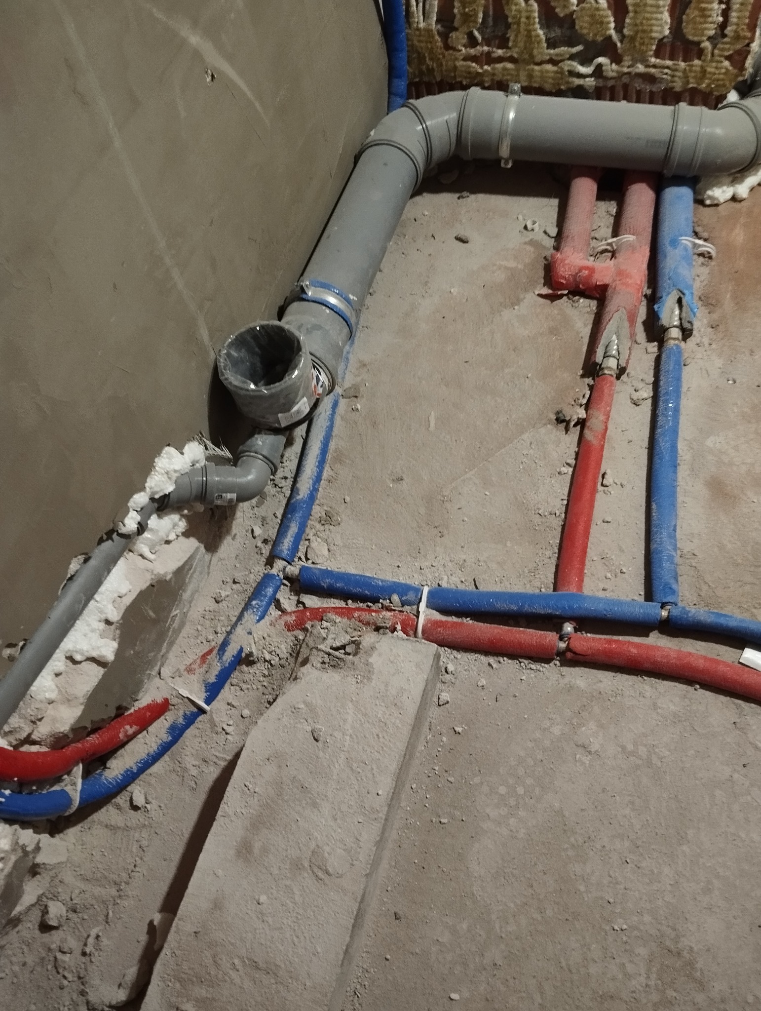 Szare rury kanalizacyjne i czerwono-niebieskie rury wodne, zamocowane do ściany i podłogi, z widoczną pianką montażową, w surowym stanie wykończenia.