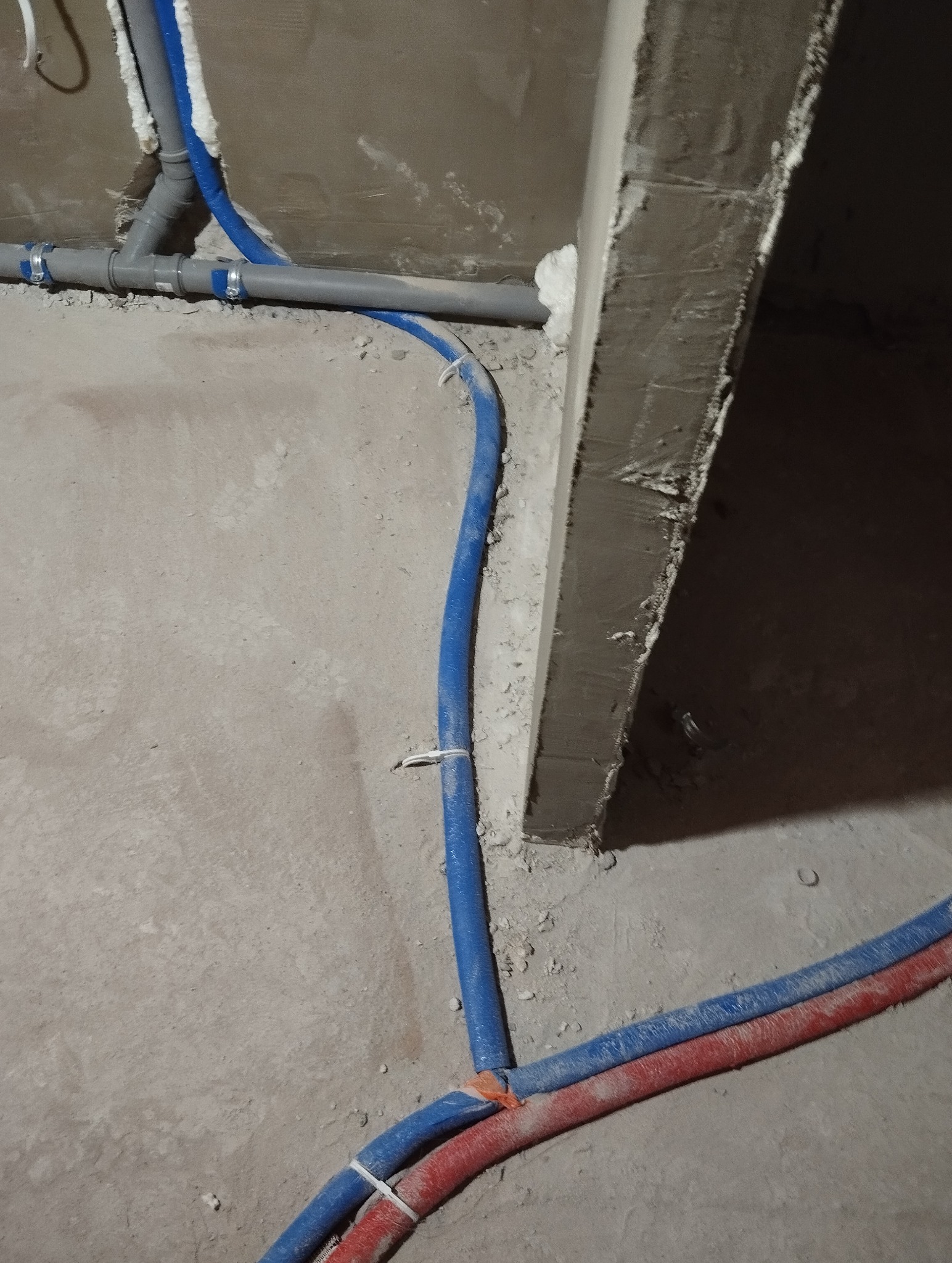 Instalacja wodna: rury w kolorze niebieskim i czerwonym ułożone na surowej posadzce w trakcie montażu, widoczne połączenia i izolacja. Ściana w tle w stanie surowym.