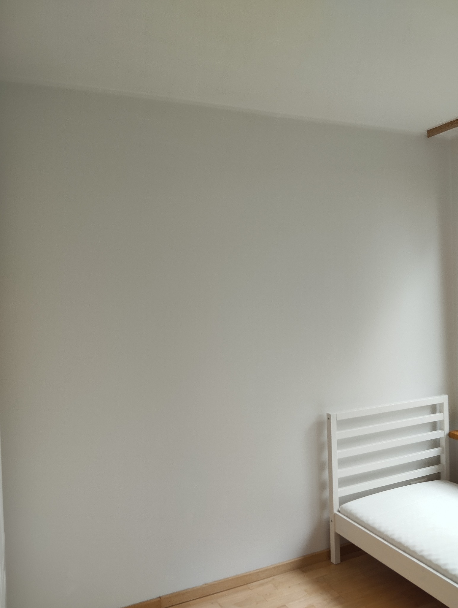 Jasny pokój po malowaniu ścian na szaro, z widocznym fragmentem białego łóżka i drewnianej podłogi. Ściana gładka, bez widocznych niedoskonałości.