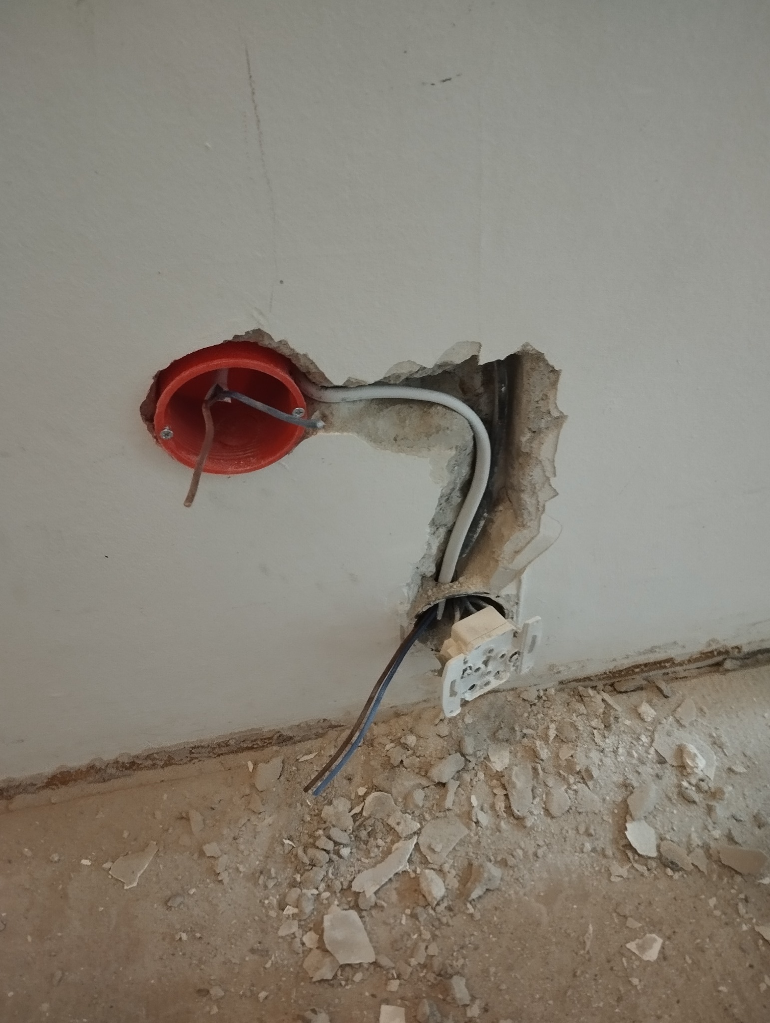 Czerwona puszka elektryczna w ścianie z wystającymi kablami i fragmentami gruzu na podłodze, widok z bliska. Prace instalacyjne w trakcie remontu.