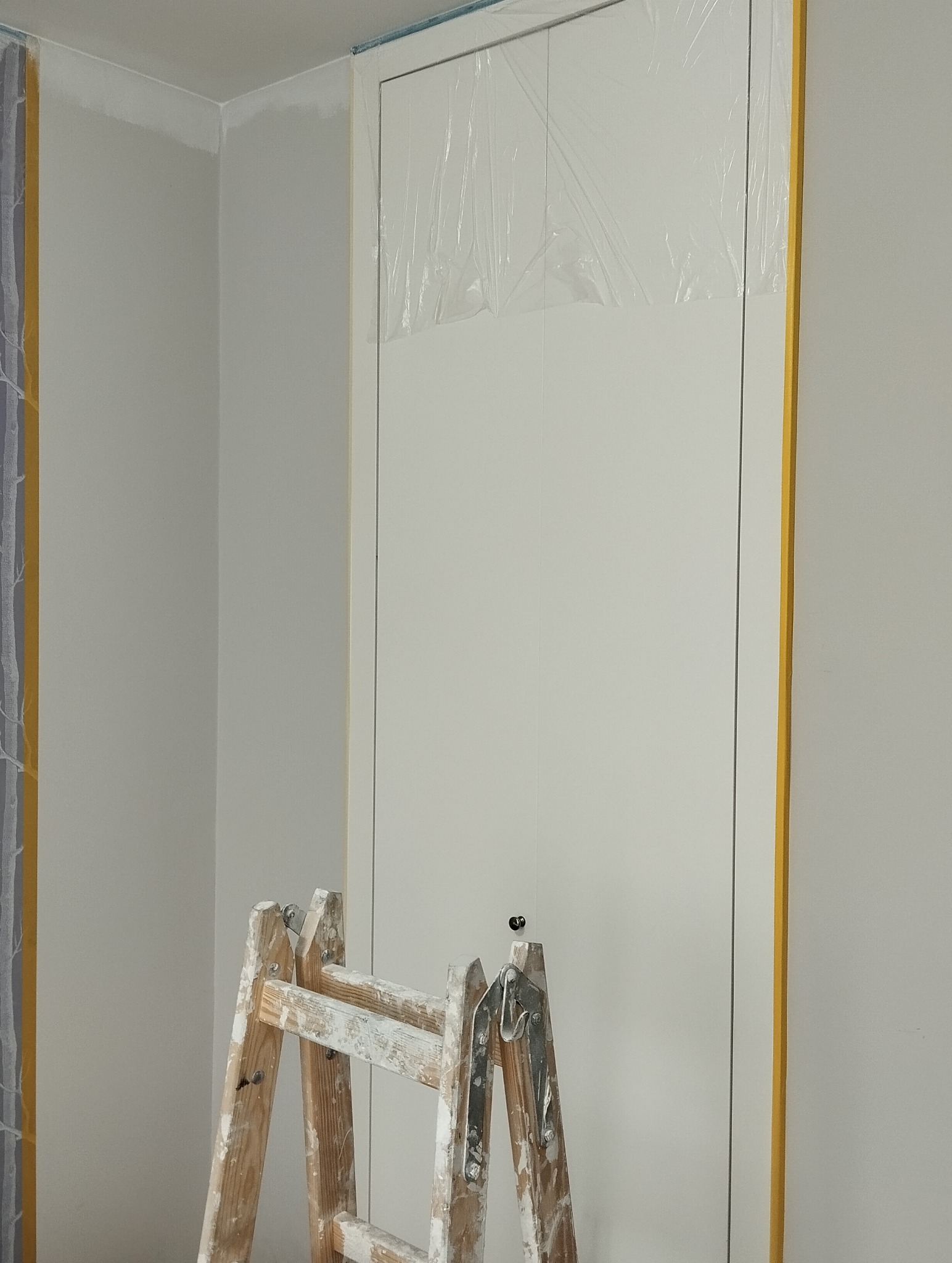 Białe drzwi zabezpieczone folią i taśmą malarską, drabina malarska w pokoju o świeżo pomalowanych ścianach na szaro. Widok z dołu, detale przygotowania do malowania.