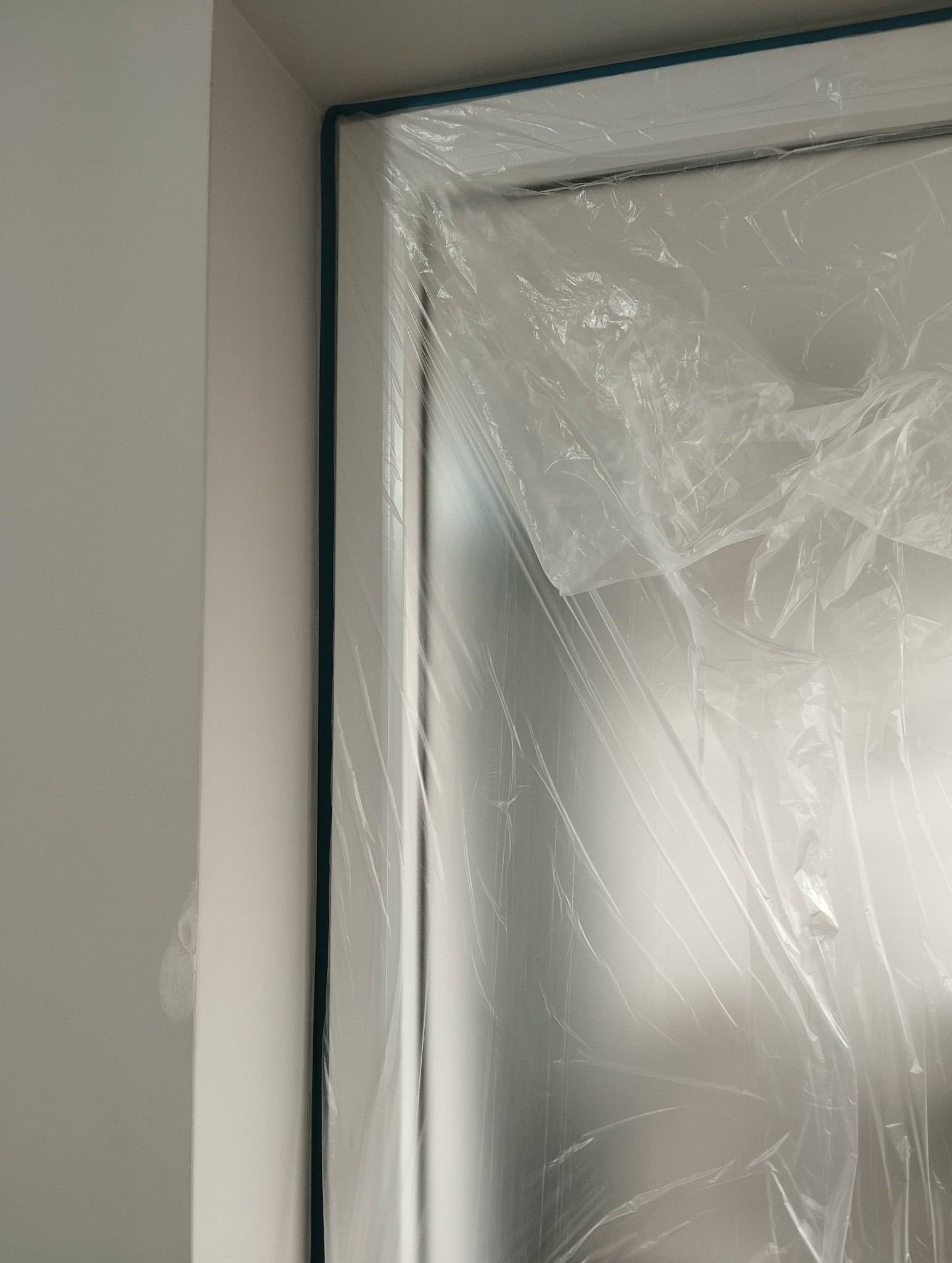 Przygotowanie do malowania: zabezpieczone folią okno z niebieską taśmą malarską, fragment ściany z widocznym punktowym ubytkiem przed malowaniem. Ochrona przed zabrudzeniem.