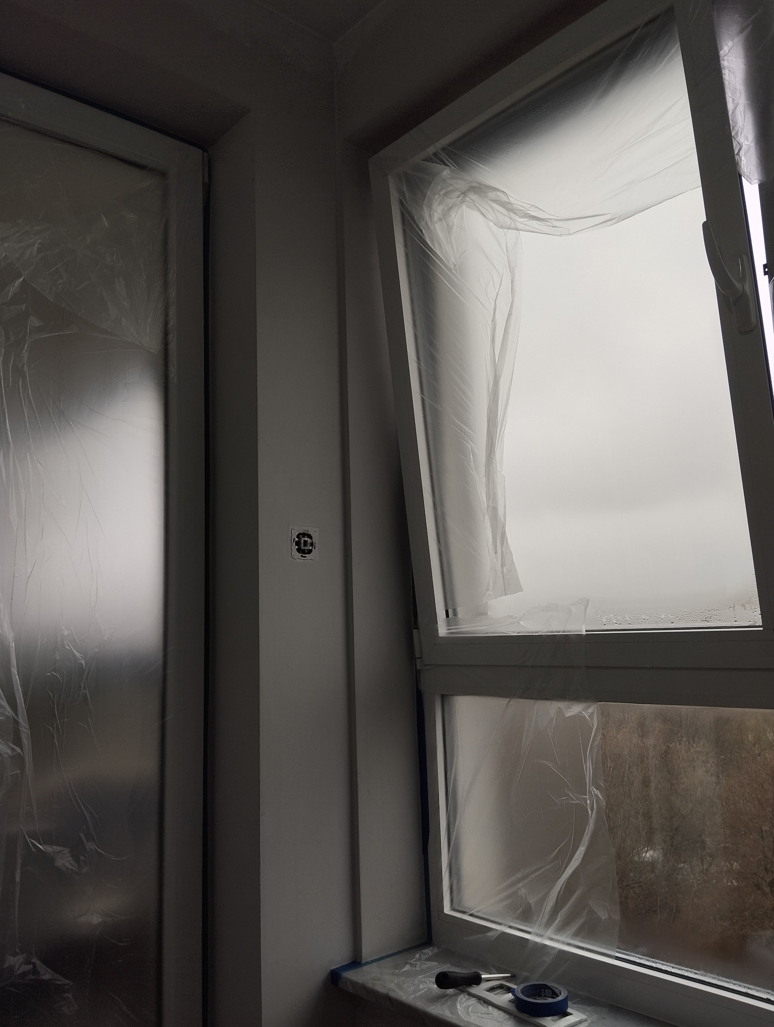 Przygotowanie do malowania: okno i drzwi zabezpieczone folią malarską w mieszkaniu w Warszawie. Widoczny fragment ściany z gniazdkiem i narzędzia malarskie.