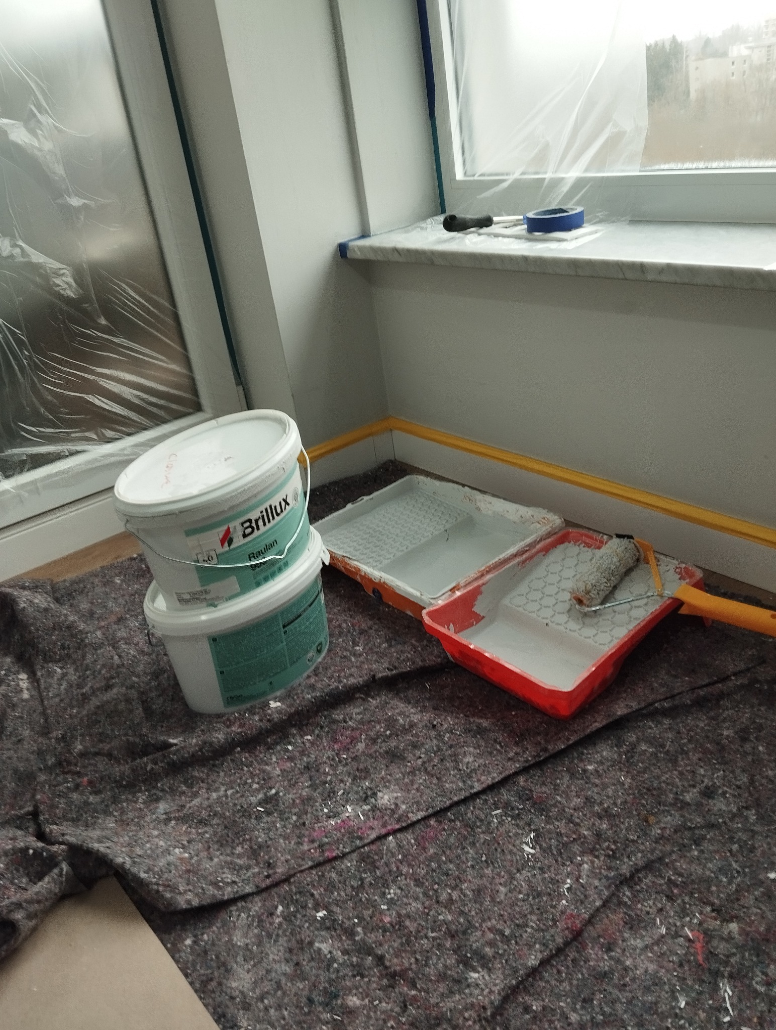 Przygotowania do malowania: wiadro farby Brillux, kuwety z farbą i wałek na podłodze zabezpieczonej folią malarską. Okno zabezpieczone folią.