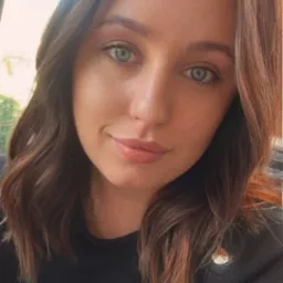 Portret kobiety o zielonych oczach i brązowych włosach, ubrana w czarną bluzkę. Uśmiech delikatny, makijaż naturalny. Oświetlenie dzienne, miękkie.