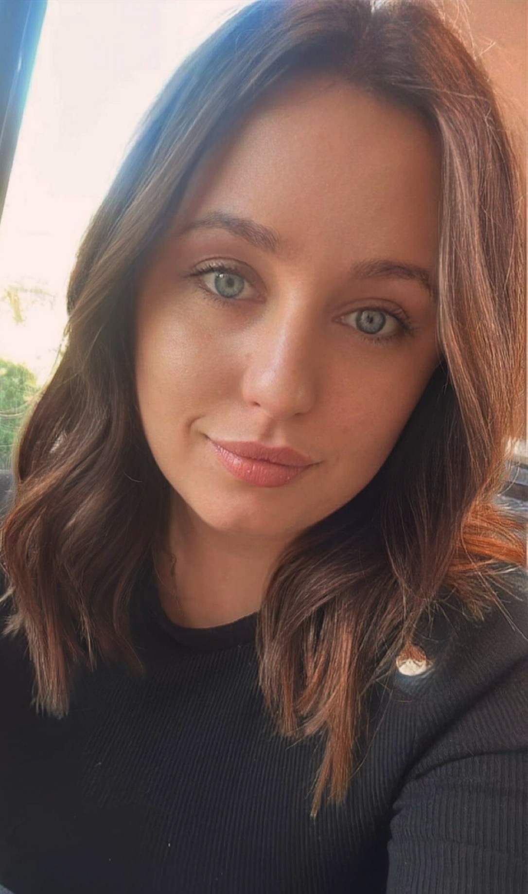 Portret kobiety o zielonych oczach i brązowych włosach, ubrana w czarną bluzkę. Uśmiech delikatny, makijaż naturalny. Oświetlenie dzienne, miękkie.