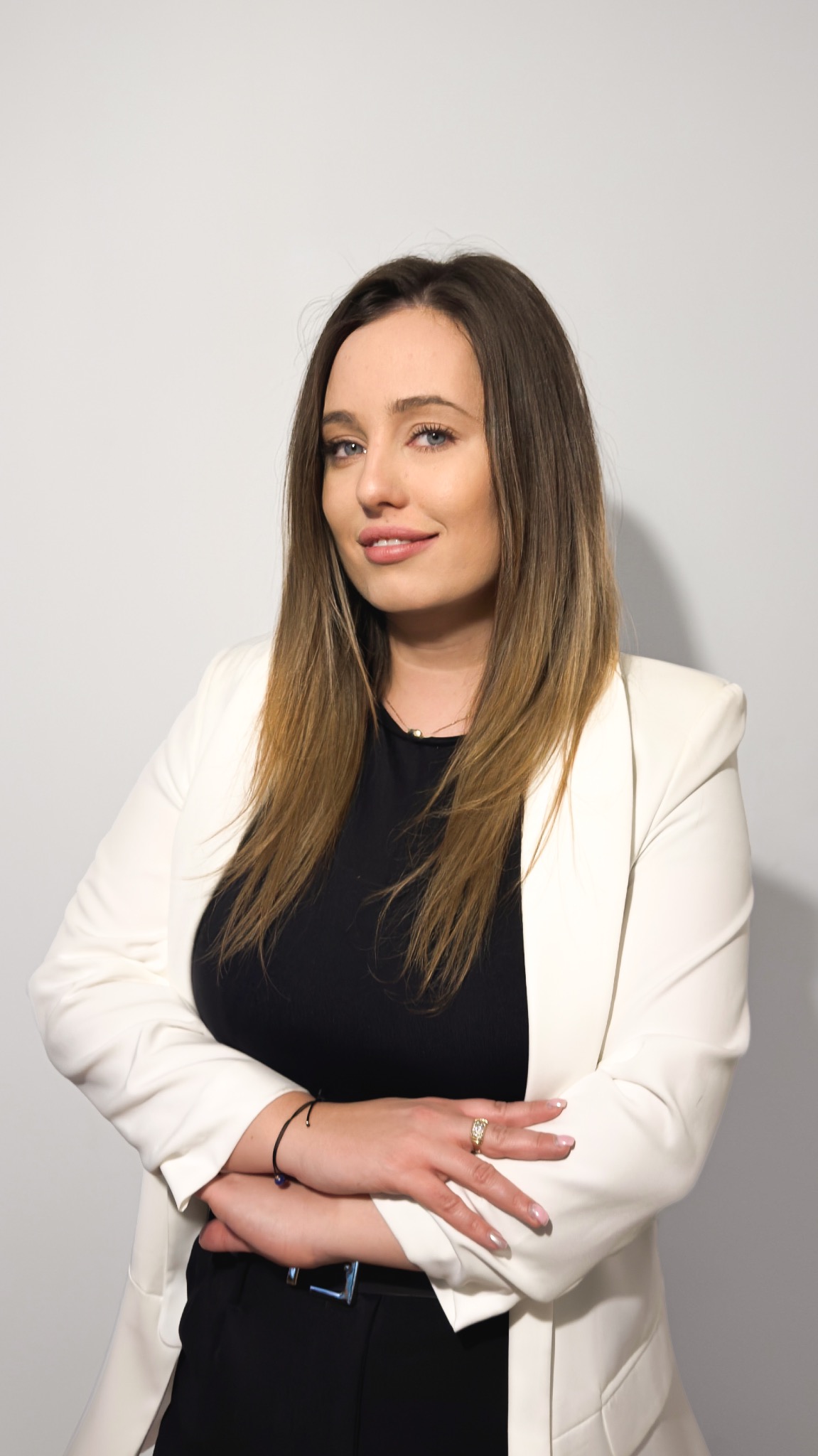 Portret kobiety w białej marynarce i czarnej bluzce, z założonymi rękami, uśmiechającej się do kamery na jasnym tle. Widoczny pierścionek i bransoletka.
