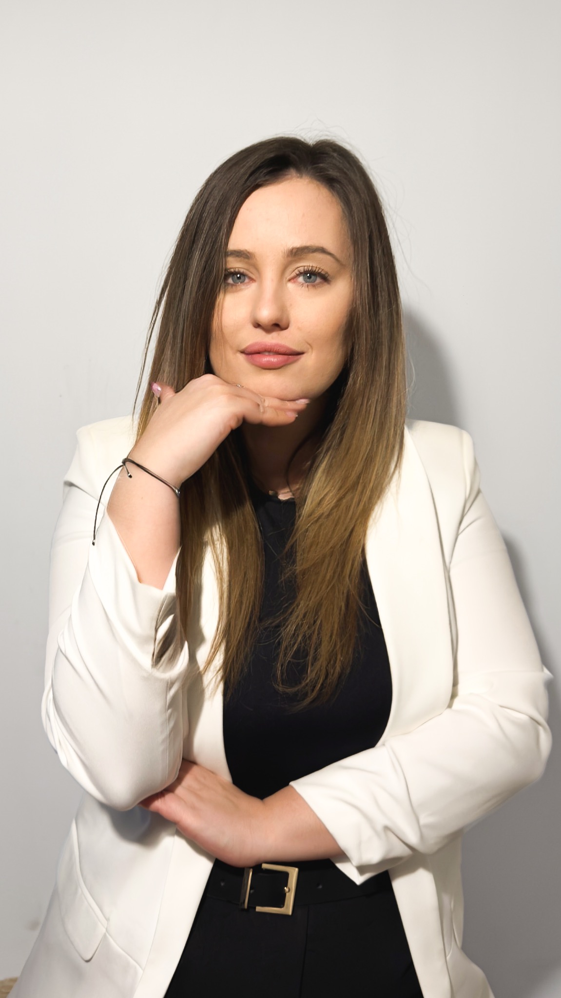 Portret kobiety w białej marynarce, z ręką pod brodą, na jasnym tle. Profesjonalny wizerunek, odpowiedni dla doradcy lub agenta.