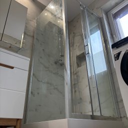 Remont4you.com.pl Dariusz Rodziewicz - Nowoczesna kabina prysznicowa z transparentnego szkła, wykończona srebrnymi profilami, osadzona na niskim, białym brodziku w łazience wyłożonej marmurkowymi płytkami, widok z dołu na kabinę...