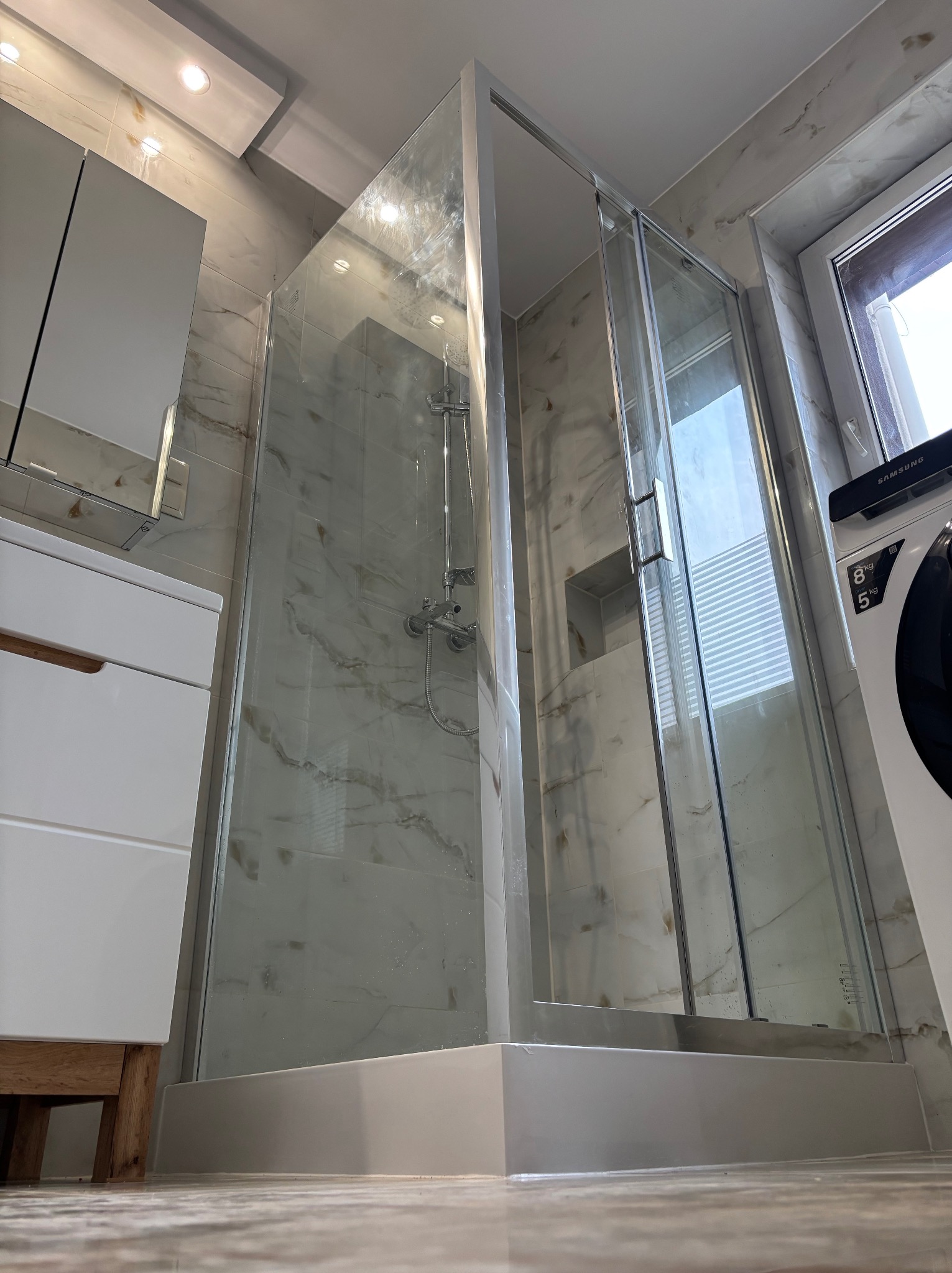 Nowoczesna kabina prysznicowa z transparentnego szkła, wykończona srebrnymi profilami, osadzona na niskim, białym brodziku w łazience wyłożonej marmurkowymi płytkami, widok z dołu na kabinę...