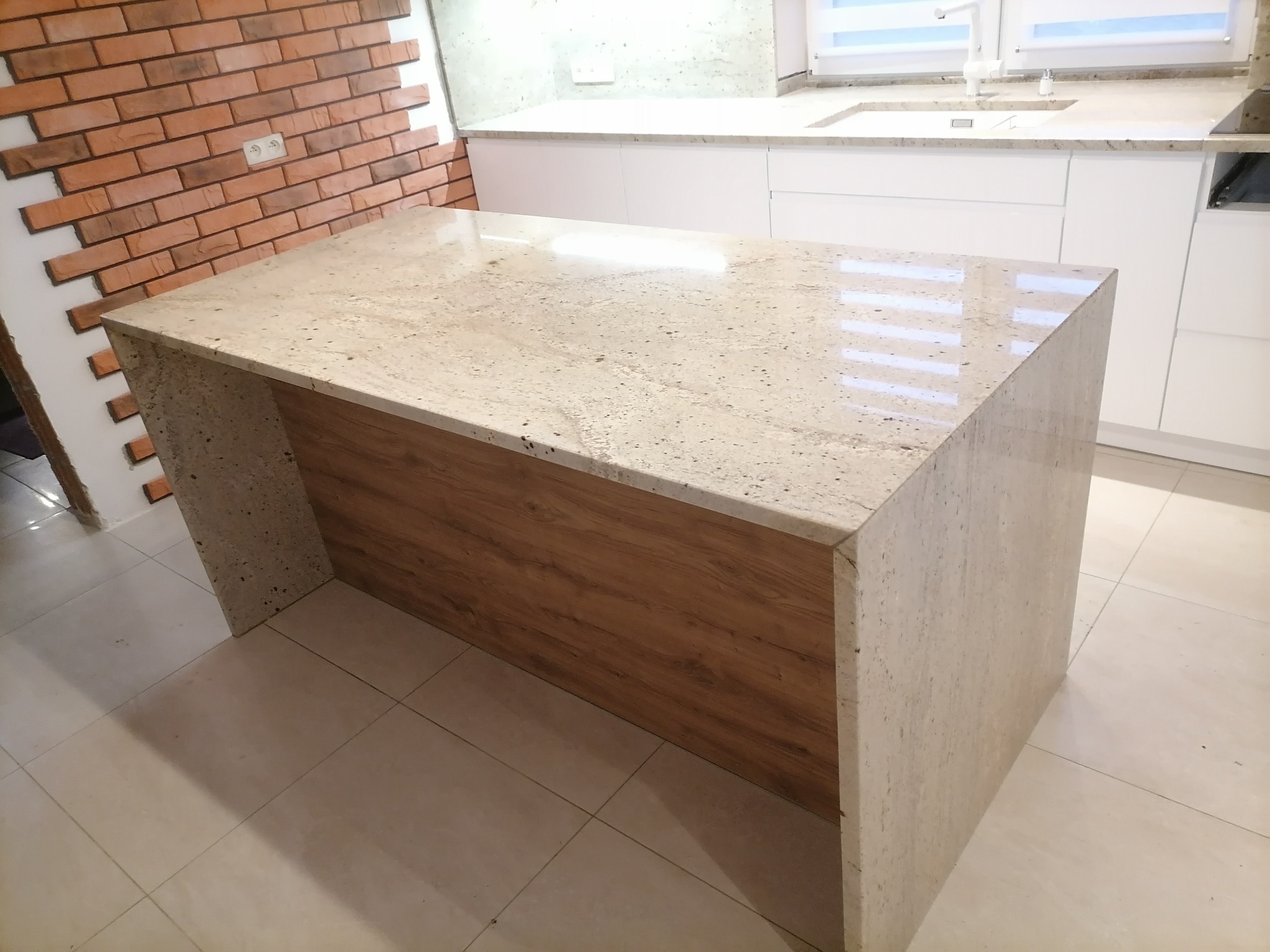 Beżowy blat kamienny z drewnianą wstawką, tworzący wyspę kuchenną w domu w Wolicy. Widoczna ceglana ściana w tle.