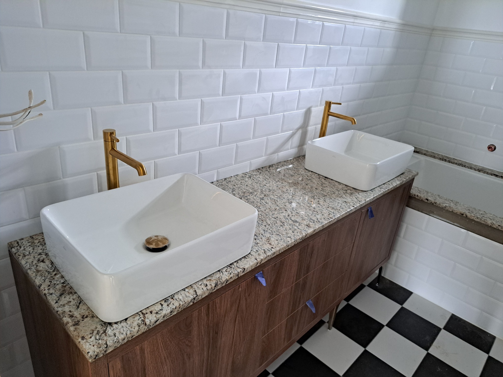 Dwie białe, prostokątne umywalki nablatowe z złotymi bateriami na blacie z jasnego konglomeratu, białe kafelki na ścianach i czarno-biała podłoga w szachownicę.