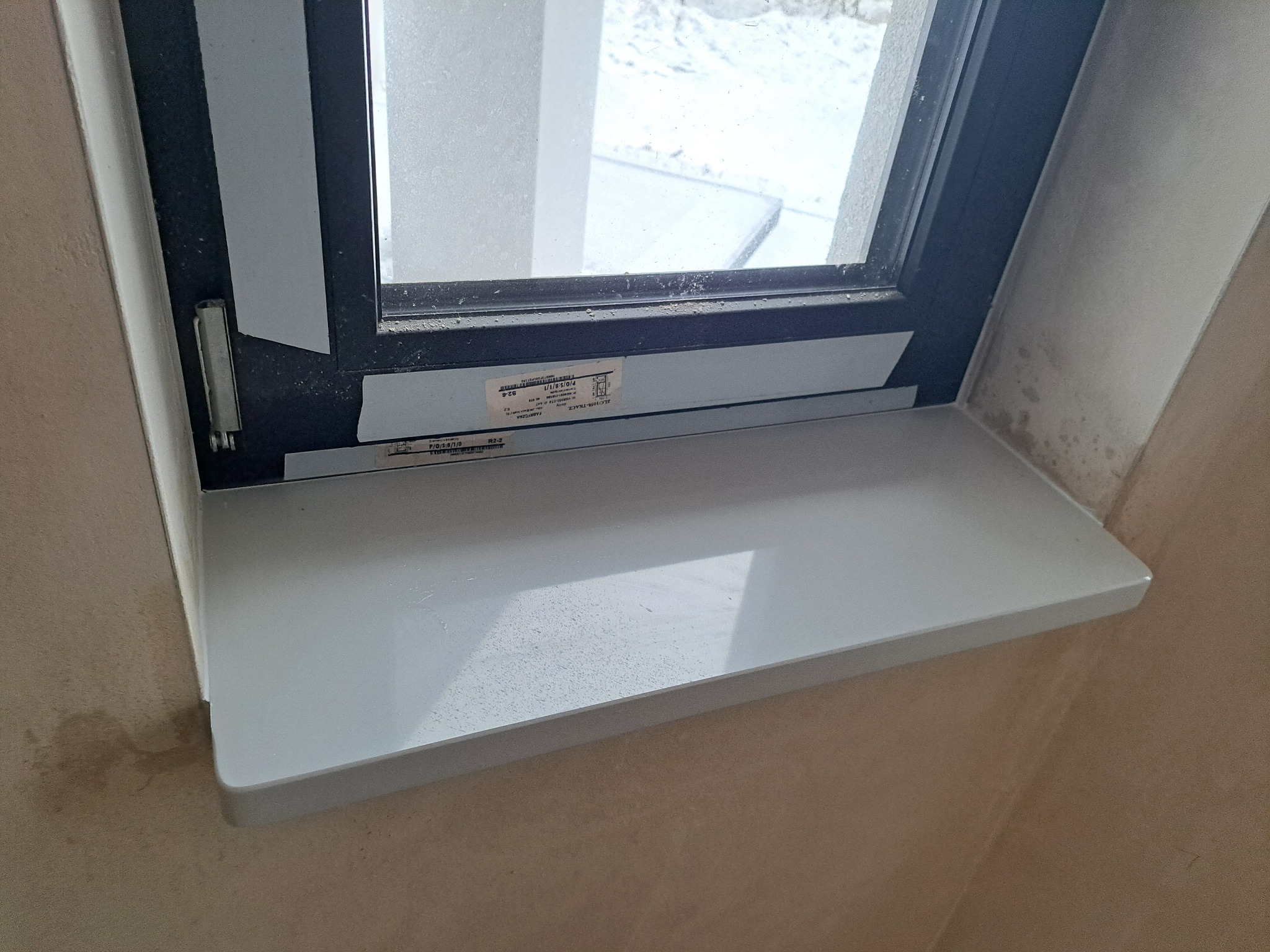 Zamontowany biały parapet z konglomeratu pod oknem z czarną ramą, widoczne zabrudzenia na parapecie i ścianie, naklejki producenta na ramie okiennej.