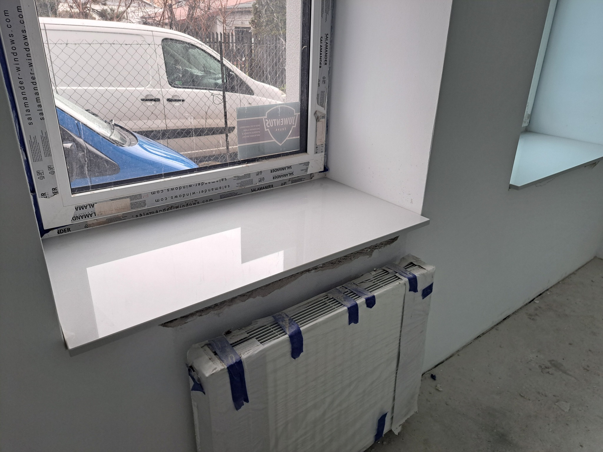 Biały, błyszczący parapet wewnętrzny z konglomeratu, zamontowany pod oknem z widocznym na zewnątrz samochodem i budynkiem, z zabezpieczonym grzejnikiem pod spodem, w trakcie prac wykończeniowych.