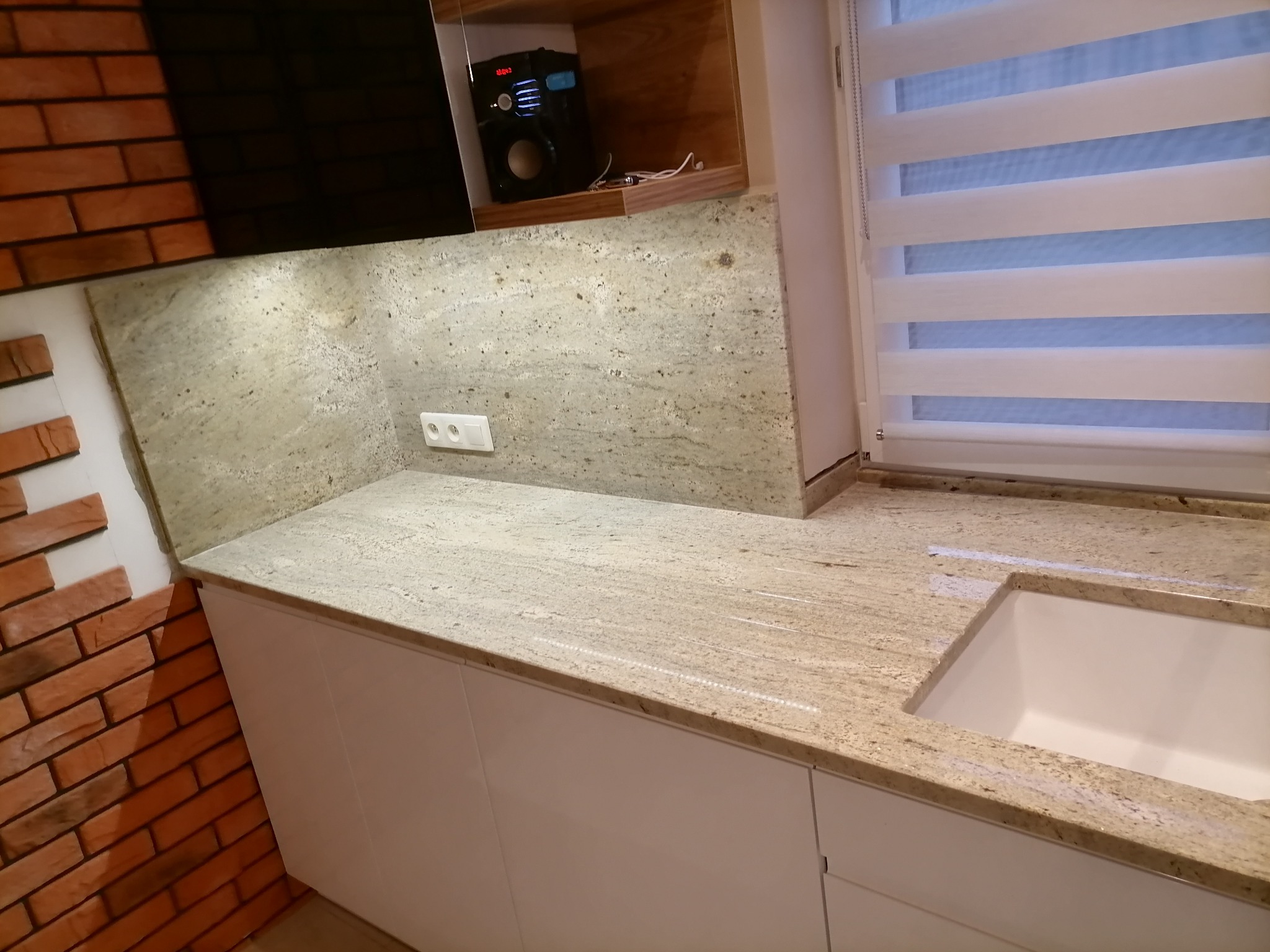 Jasny blat z kamienia naturalnego z wyciętym otworem na zlew, białe fronty szafek, ceglana ściana w tle, roleta okienna w paski.