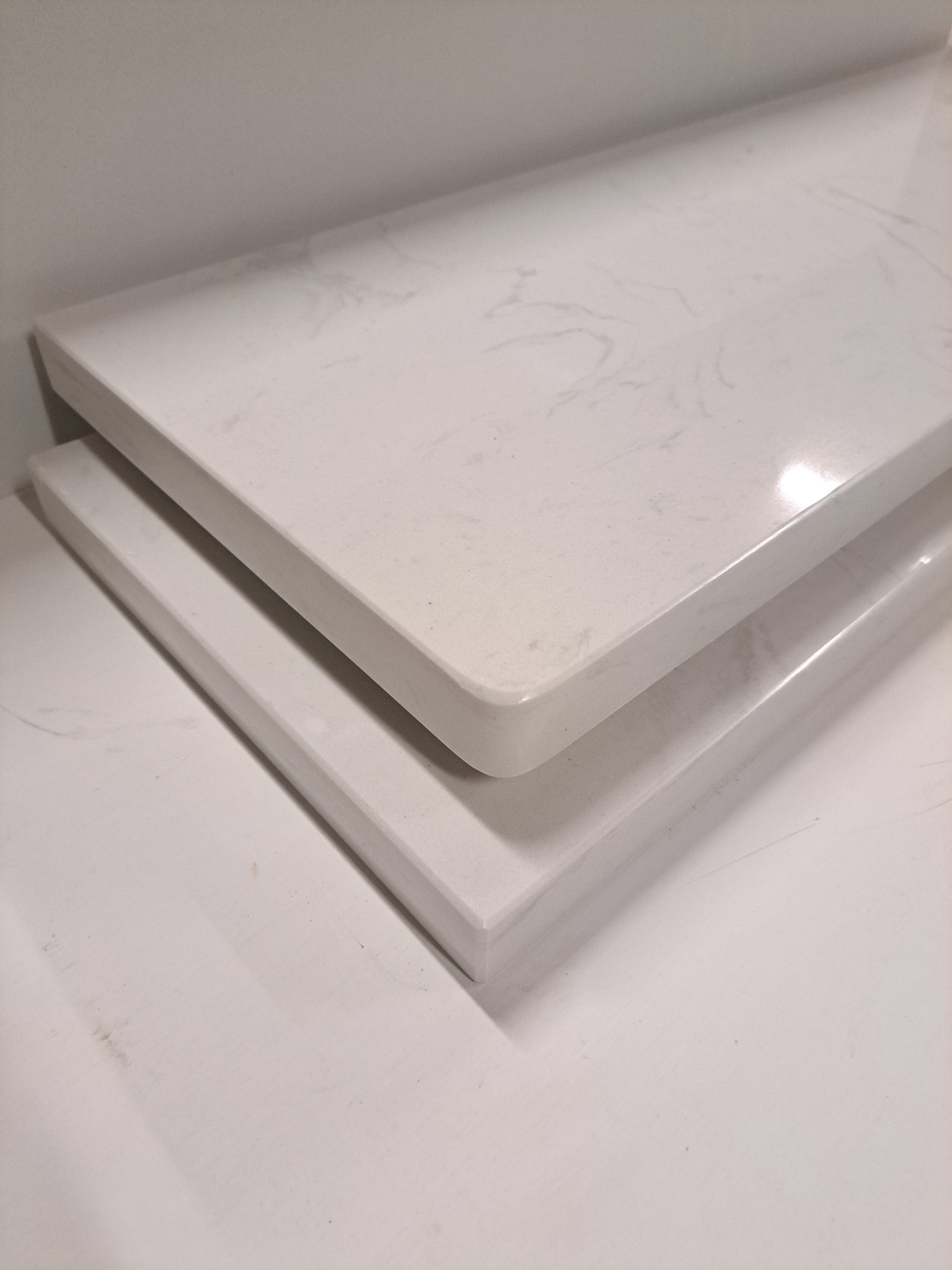 Dwa białe, marmurkowe elementy z zaokrąglonymi krawędziami, prawdopodobnie fragmenty blatów lub parapetów, ułożone jeden na drugim.