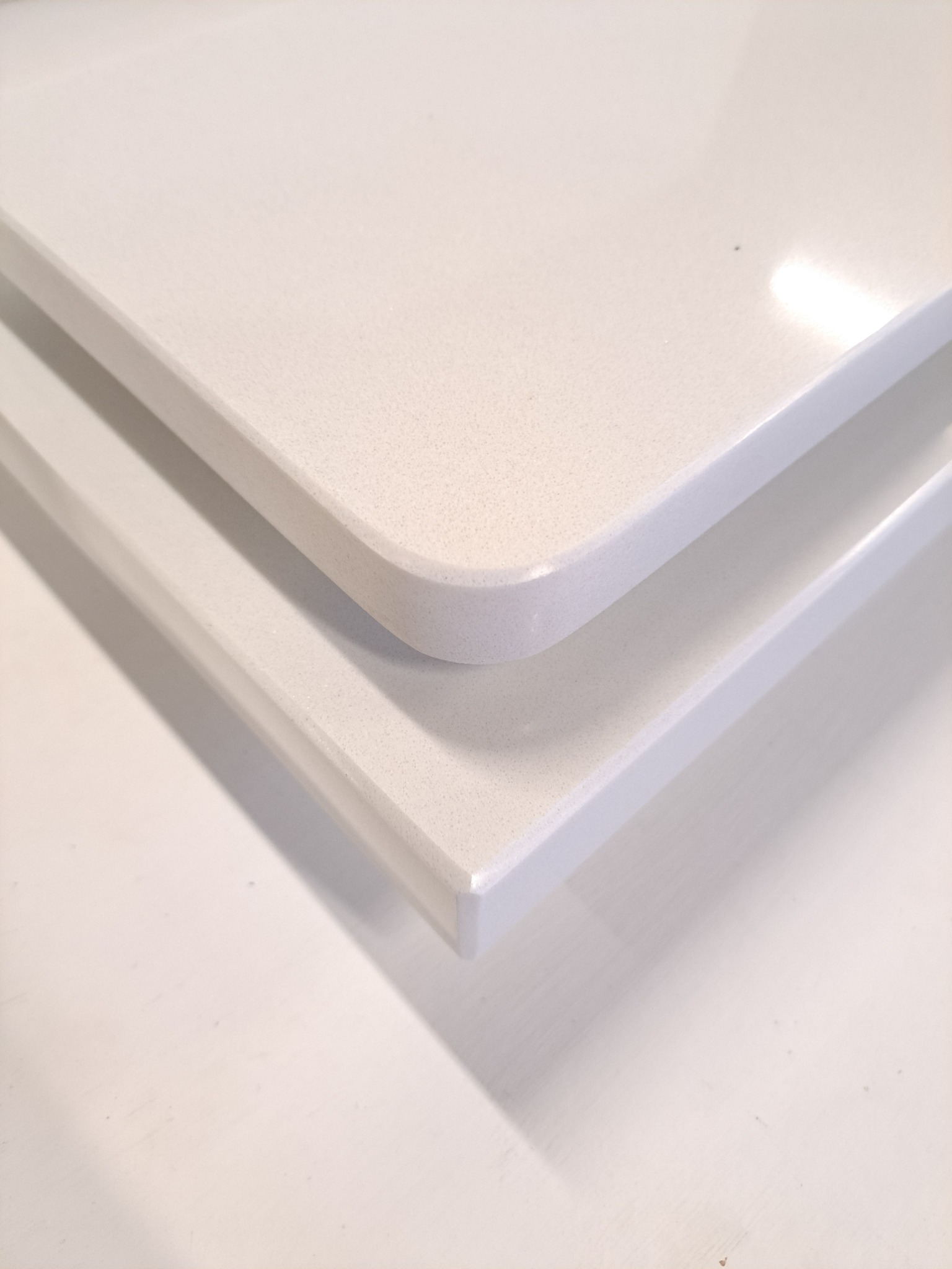 Dwa białe blaty z konglomeratu kwarcowego, jeden z zaokrąglonym narożnikiem, ułożone jeden na drugim, widoczne z bliska w neutralnym oświetleniu.